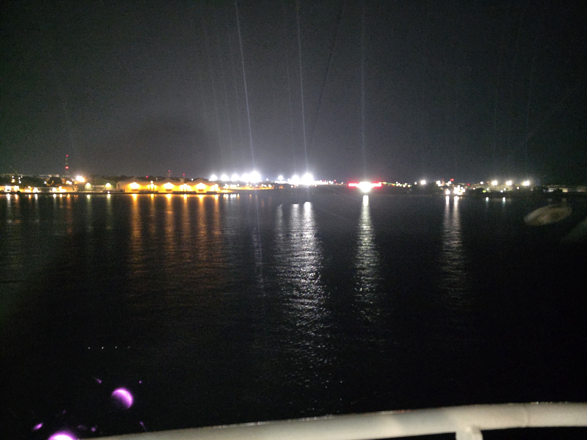 The image shows a night view of a waterfront in Brindisi, Italy. The scene is dominated by the dark water of the harbor reflecting the lights of buildings and structures along the shoreline. The lights are primarily orange and white, with some red lights visible on what appears to be a distant tower. The reflections on the water are elongated and distorted due to the movement of the water and the low light conditions. In the foreground, a white railing is partially visible, suggesting the vantage point is from a boat or a dock. There are no people or discernible objects in the water itself, except for some faint, blurry purple lights in the lower left corner, which might be from the same vessel the photo is taken from. The overall atmosphere is dark and industrial, typical of a port city at night.