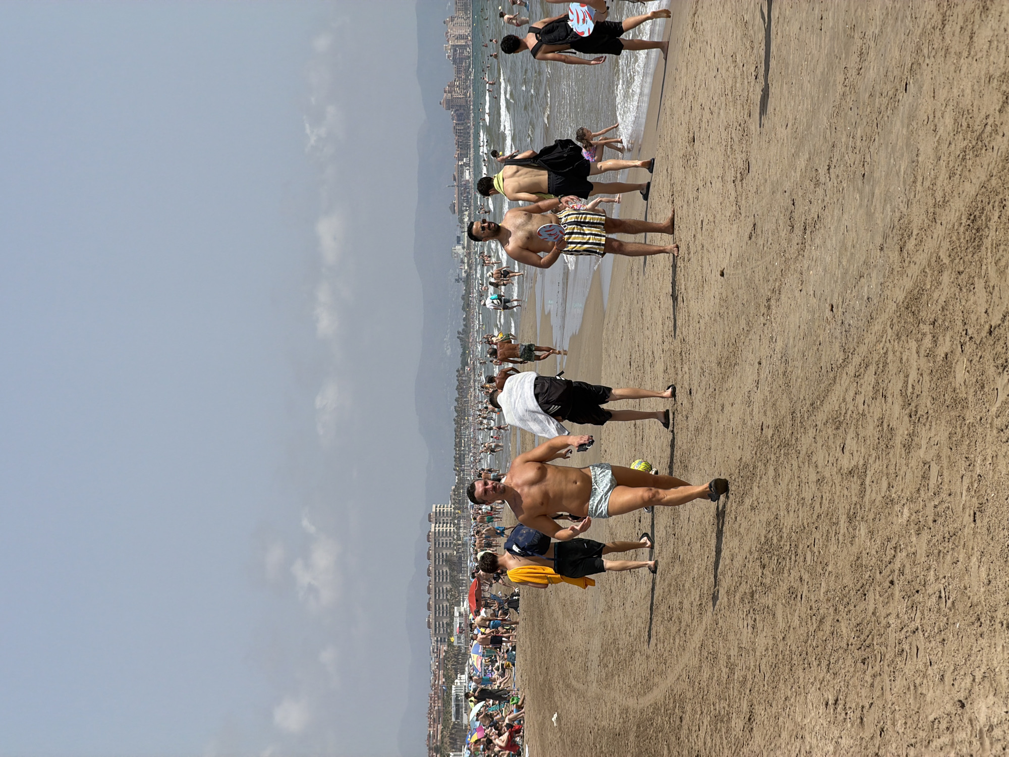 An outdoor, eye-level, full shot captures a sandy beach with numerous people. The sand is packed and displays tire tracks. In the foreground, a man with a muscular physique walks towards the camera wearing light blue swim trunks. He has his arms slightly outstretched. Behind him, to his left, another man in black shorts and a yellow towel draped over his shoulder walks away from the camera. To the right of the foreground man, a man in striped swim trunks, sunglasses, and a yellow towel around his neck holds a paddle. Further back, other beachgoers are visible, including individuals swimming in the ocean, sunbathing under umbrellas, and walking along the shoreline. The background features a hazy cityscape with buildings nestled against rolling hills under a bright, overcast sky. The ocean is calm with gentle waves.