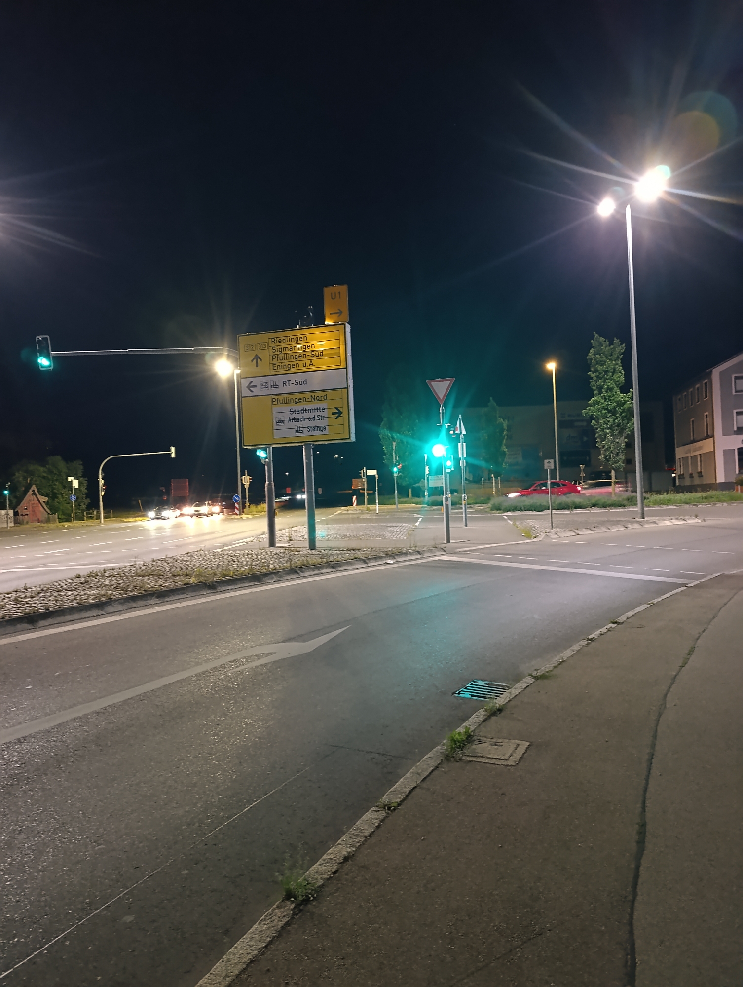 This detailed nighttime image captures a multi-lane road intersection in Pfullingen, Germany, specifically the junction of Sigmaringer Straße (B312) and Arbachstraße / Gammertinger Straße (L387). The scene is illuminated by numerous streetlights, with one on the right emitting significant lens flares against the dark sky.
In the immediate foreground, an asphalt road surface displays clear white lane markings, including a solid white line on the left and a prominent right-turn arrow. A concrete curb delineates the edge of the roadway from a paved sidewalk on the right, where a metal drainage grate is visible.
At the center-left, a prominent yellow, multi-panel directional sign is mounted on a pole. The top panel, marked with "312 / 313," directs "Riedlingen, Sigmaringen, Pfullingen-Süd, Eningen u.A." with an upward arrow. The middle panel features a left arrow for "RT-Süd," while the bottom panel indicates "Pfullingen-Nord, Stadtmitte, Arbach-o.d.Str, Steinge" with a right arrow. Above this main sign, a smaller yellow "U1" sign with a right arrow is attached.
Multiple traffic light signals are visible, all displaying green. A horizontal traffic light spans the leftmost lane, and a vertical traffic light pole stands closer to the main sign. Further into the intersection on the right, a red triangular yield sign with a white background is present. An unpaved median strip with sparse vegetation is situated in the center of the intersection.
Several vehicles are observed. A red passenger car is in motion through the intersection on the right, evidenced by the motion blur on its taillights. A darker vehicle follows it. In the distance, multiple vehicles with illuminated headlights are visible, some appearing stationary or moving slowly.
The background on the far left includes a reddish-brown building with a distinctive roofline, while further right are areas of vegetation and trees. On the right side of the image, multi-story buildings, consistent with residential or commercial structures, are illuminated by the ambient light. No individuals are visible within the frame.