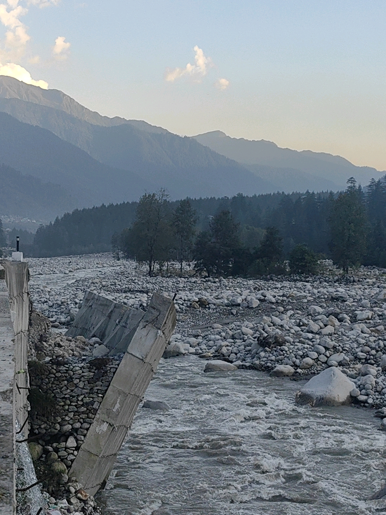 In Vashisht, India, a damaged concrete structure, possibly part of a bridge or road, lies adjacent to a rocky riverbed. The structure's concrete segments are broken and tilted, with exposed rebar. The river flows with murky water, interspersed with numerous large, grey rocks. Beyond the river, a dense forest of dark green trees stretches across the landscape, backed by layered, misty mountains that fade into the hazy sky. The sky is a pale blue with scattered white clouds. The scene suggests a natural disaster or severe weather event that has caused significant damage to man-made infrastructure and altered the river's course. There are no people or animals visible in the frame.