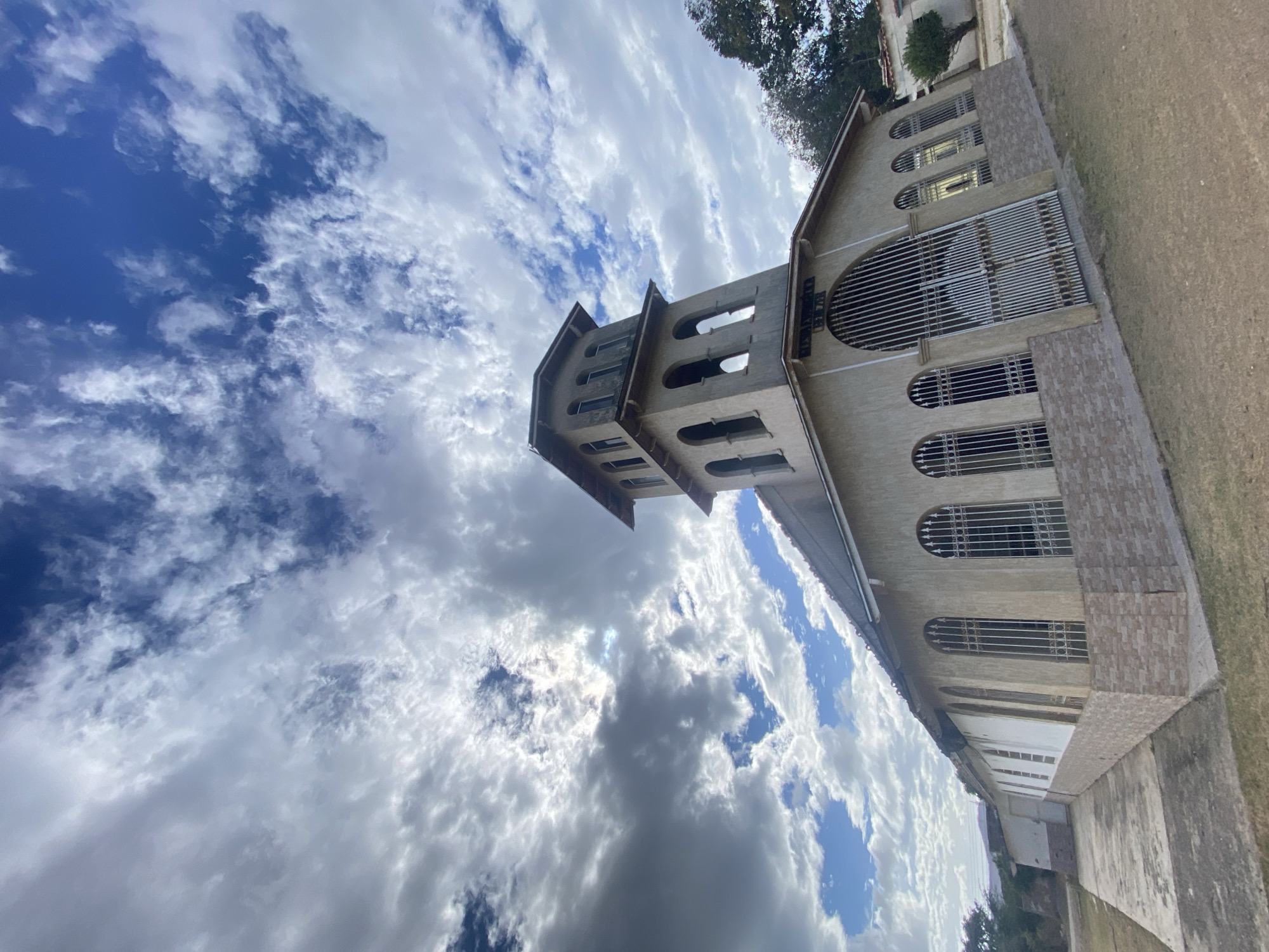 The image shows the Basilica Menor Del Cristo Del Buen Viaje, a minor basilica located in Juan Griego, Venezuela. The building features a tall, multi-tiered bell tower with arched openings, and a facade with arched windows covered by metal bars. The exterior of the basilica is light-colored. The sky is filled with white and gray clouds, and a patch of blue sky is visible. Vegetation, possibly trees, can be seen near the building. The photograph appears to have been taken from a low angle.