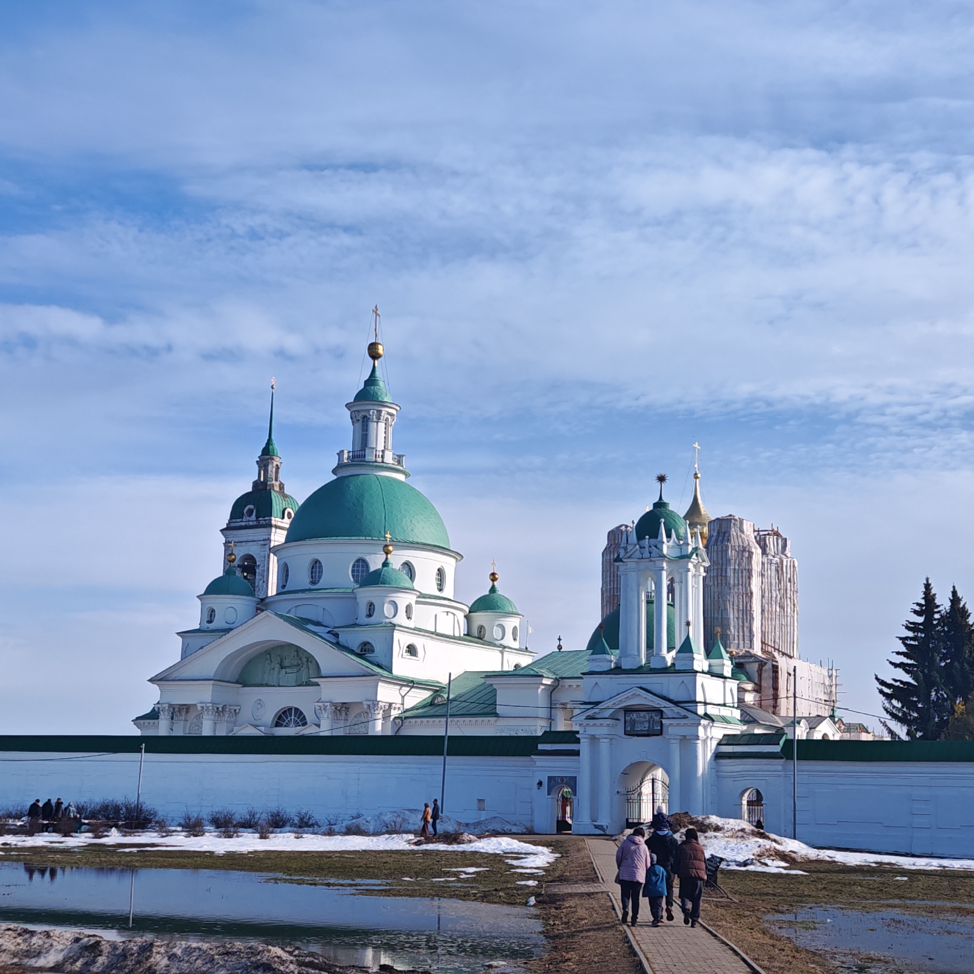 The image displays an exterior view of the Spaso-Yakovlevsky Monastery located in Rostov, Russia (Rostov Veliky, Yaroslavl Oblast). The scene is captured on a day with a partially cloudy blue sky, indicating late winter or early spring conditions with visible snowmelt. The monastery complex features prominently, characterized by white masonry structures and numerous onion-shaped domes, primarily painted green, culminating in golden spires topped with Orthodox crosses. The main cathedral, positioned centrally, exhibits a large central green dome flanked by several smaller green domes and tall spires. To its right, a significant structure is undergoing renovation, encased in white protective sheeting and scaffolding, with a golden spire discernibly extending from its top. A sturdy white masonry wall with a dark green coping encloses the complex, with a decorative arched gatehouse, adorned with columns and a small green dome, serving as an entry point on the right side of the wall.
In the immediate foreground, a broad puddle of standing water reflects the sky, bordering areas of patchy brown grass and residual snow. A narrow, unpaved path extends from the lower right towards the monastery gate. Four individuals walk along this path, observed from behind. One person wears a light-colored jacket, another a dark jacket, a child wears a blue jacket, and a fourth is in a dark vest. Further to the left, adjacent to the monastery wall, a smaller group of approximately three to four people is visible standing amidst the snow-dusted ground and sparse bushes. On the far right, outside the monastery walls, several tall, dark green coniferous trees stand against the sky.