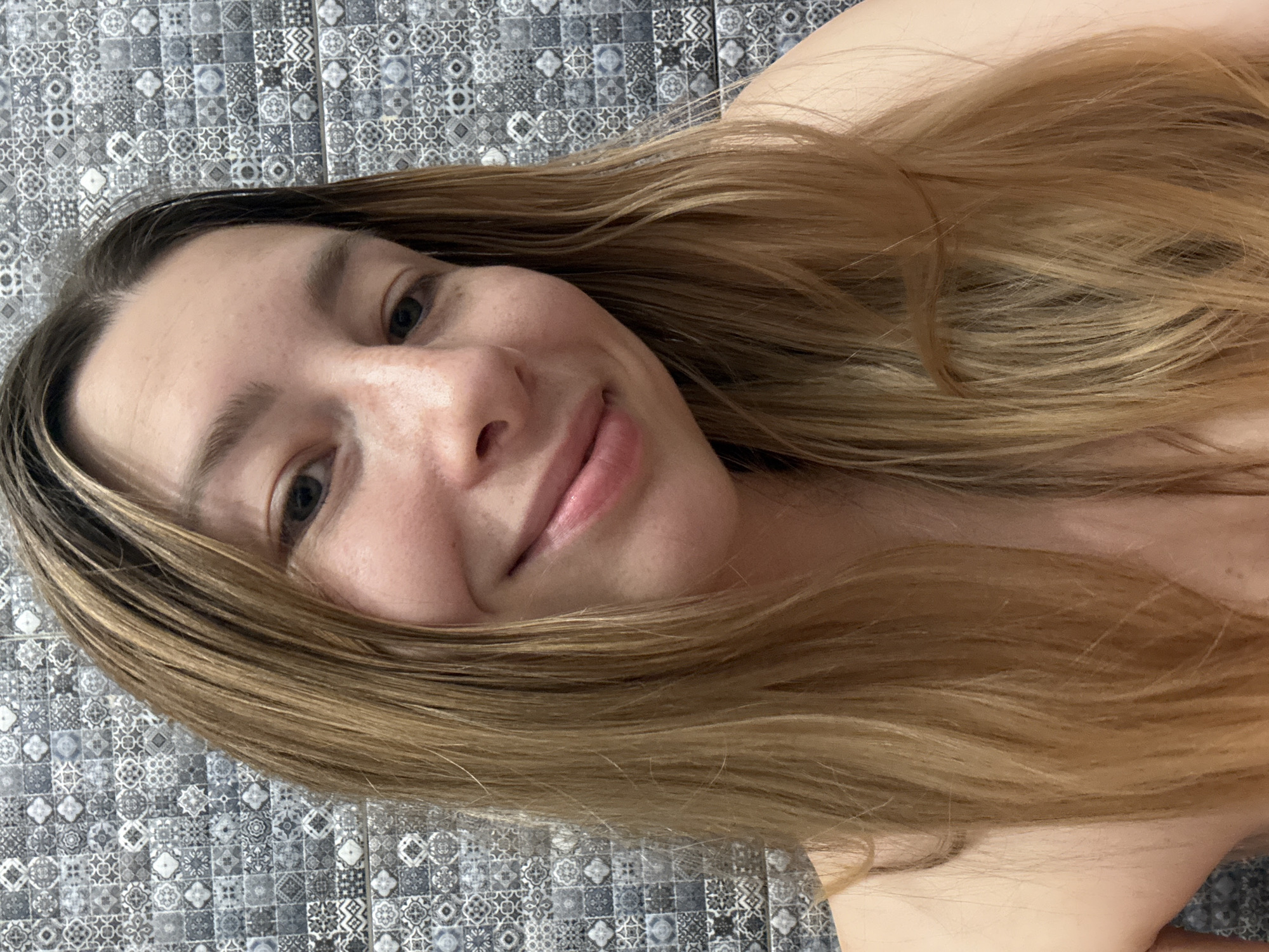 A selfie of a woman with long blonde hair is angled against a gray-patterned tile wall. Her face is visible, displaying light skin, freckles, and a subtle smile. No information suggests the location is in Moscow, Russia.