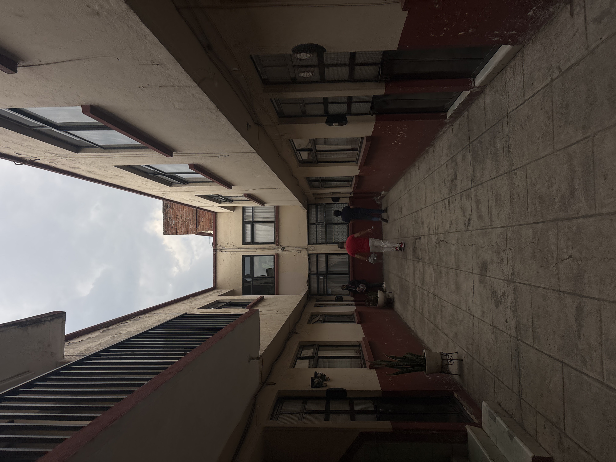 The image depicts an outdoor space between two buildings, likely an alley or courtyard. The buildings are multi-storied, with a beige color scheme and red trim. Windows are visible on both buildings. A staircase is on one side. The ground is paved with large, square tiles. Two people are in the middle of the space. One individual is wearing a dark-colored shirt and pants, while the other is wearing a red shirt and white pants. The scene appears to be in Puebla, Mexico.