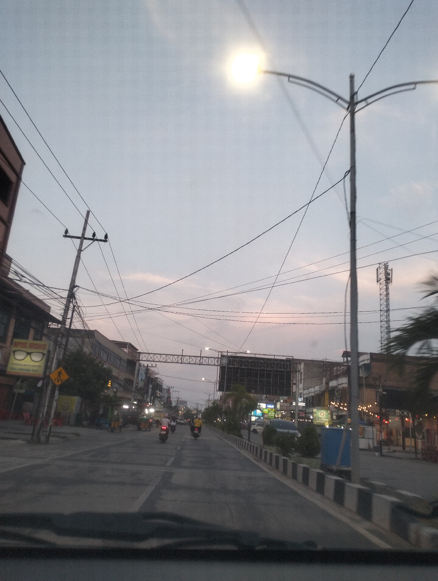 The image shows a street scene at dusk in Dumai City, Indonesia. The sky is a pale blue with some pinkish hues, indicating the sun has recently set or is about to rise. A bright street lamp is illuminated on the right side of the frame.
The street is paved and has clear lane markings. There are several buildings lining the street on both sides, appearing to be shops or commercial establishments. Some of these buildings have signs, and lights are visible in some windows and from decorative string lights along the storefronts. A tall cellular tower is visible in the distance on the right.
Several motorbikes are on the road. Two are in the foreground, moving away from the camera, and others are further down the street. There are power lines and utility poles crisscrossing the sky above the street. A traffic sign showing a pedestrian crossing is visible on the left. The overall impression is a typical urban street in Southeast Asia as evening transitions into night.