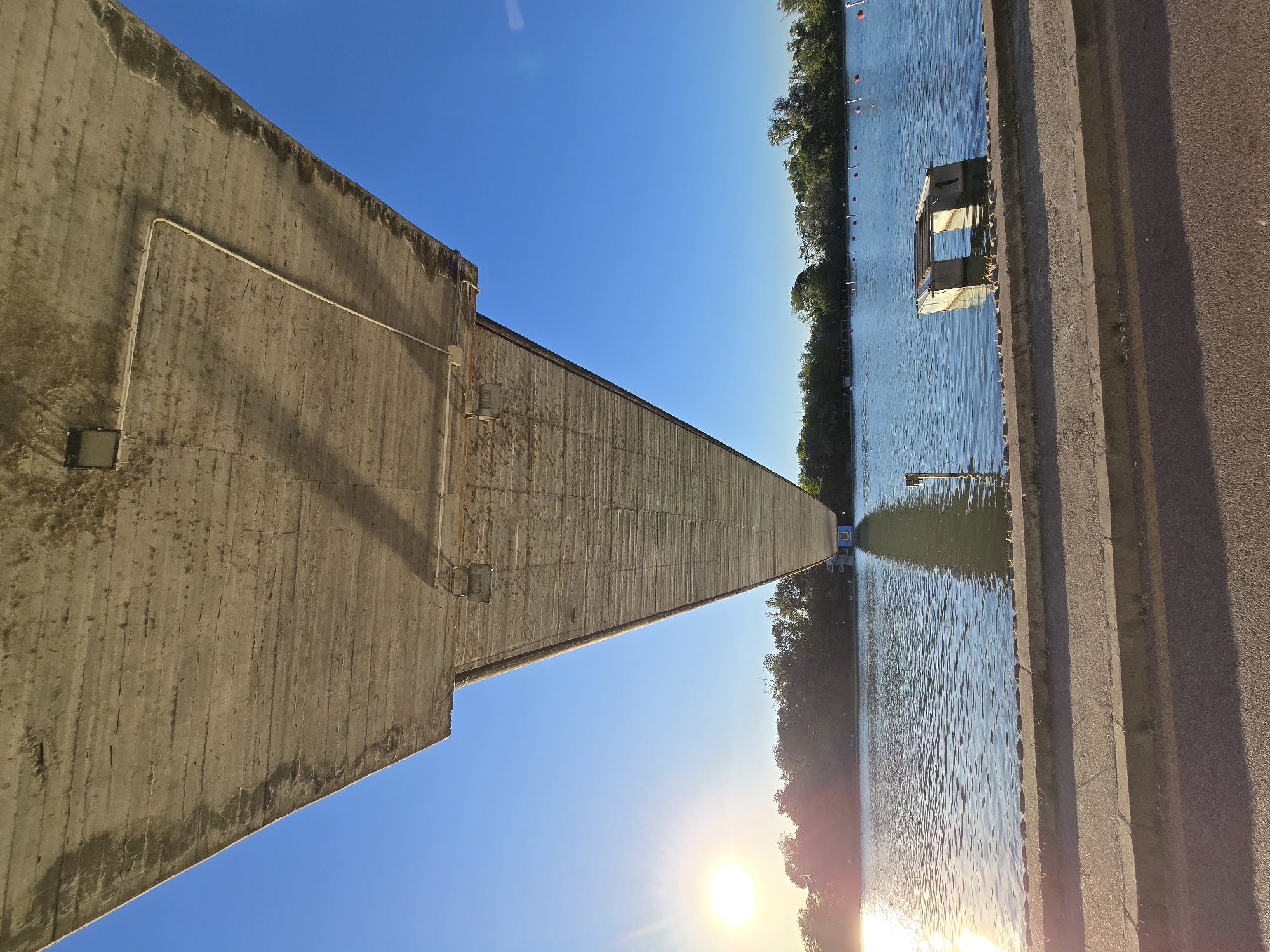 Here is a description of the image:
The image shows a landscape featuring a concrete structure alongside a body of water, likely a river or lake, under a clear blue sky. A low building is visible in the water, with greenery lining the far shore. The sun shines brightly, reflecting off the water's surface. A sidewalk or road runs parallel to the water. The photo appears to be taken from a low angle, possibly at ground level.