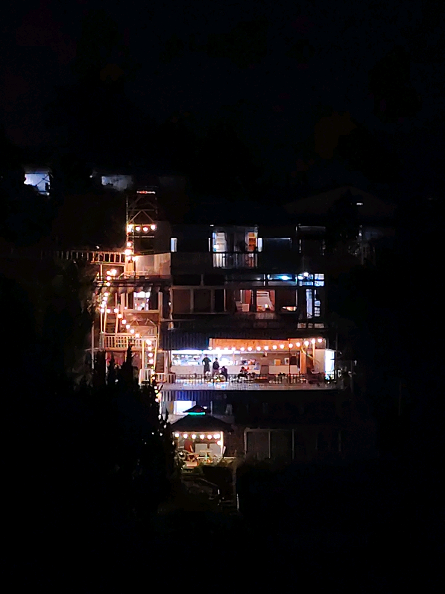 The image captures a multi-story building illuminated at night in Da Lat, Vietnam. The building features a rustic, layered architecture with several balconies and open-air seating areas. String lights are prominently displayed along a staircase and on a lower level patio, casting a warm glow. On the lower level, several individuals are seated at tables, suggesting a dining or social gathering. The surrounding environment is dark, with only the building and its lights being visible, implying the shot was taken from a distance. The overall scene conveys a cozy and inviting atmosphere despite the darkness.
