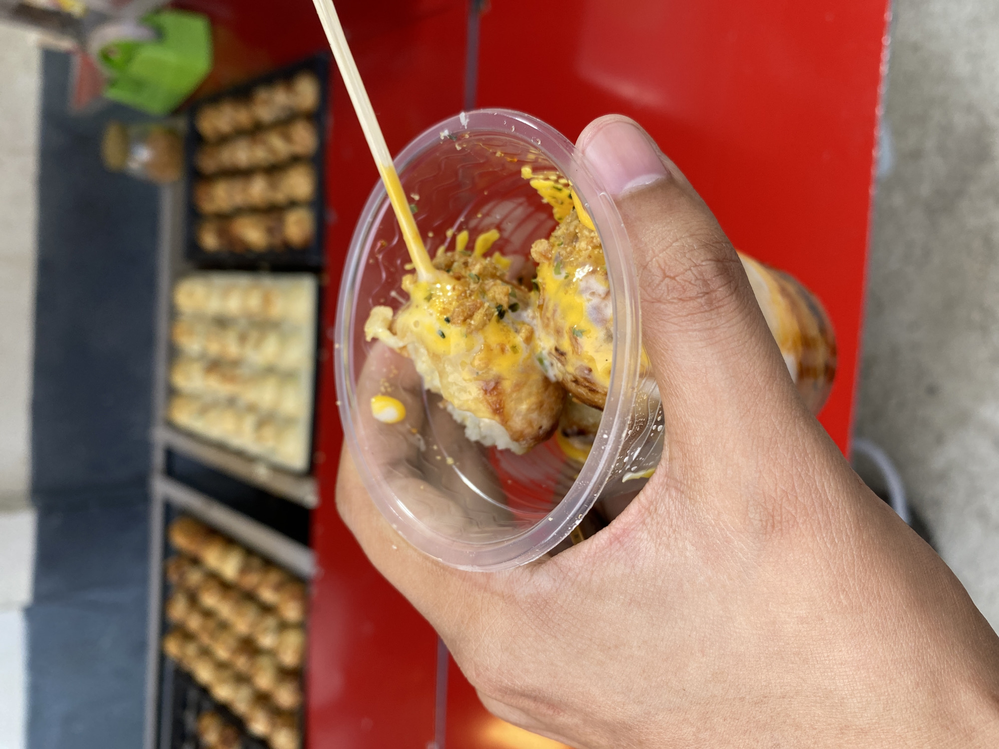 A first-person perspective shot shows a hand holding a clear plastic cup filled with takoyaki, a Japanese street food dish. The takoyaki balls are topped with a yellow sauce, crispy bits, and green specks, and one ball is skewered on a wooden stick. In the blurred background, a red cart displays trays of takoyaki being cooked. The setting appears to be an outdoor food stall. Based on visual cues such as the style of the food stall and the general aesthetic, this scene is likely from a food street or market in Davao City, Philippines, known for its vibrant street food culture.