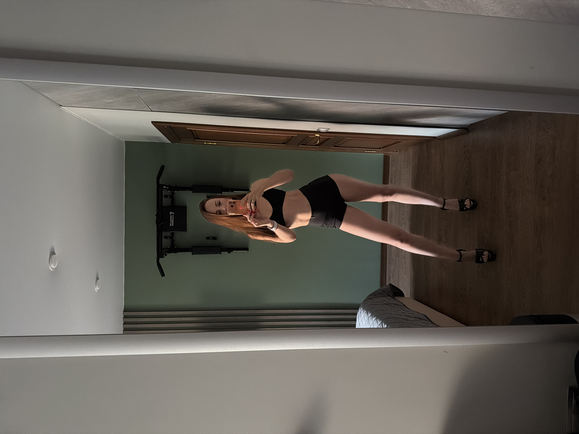 A woman with long, reddish-brown hair is taking a mirror selfie in what appears to be a bedroom. She is wearing a black sports bra and black shorts, with high-heeled sandals on her feet. In the background, a pull-up bar is mounted on a green wall, and a portion of a bed is visible. The scene is lit by natural light. The image does not provide any information about the location in Birobidzhan, Russia.