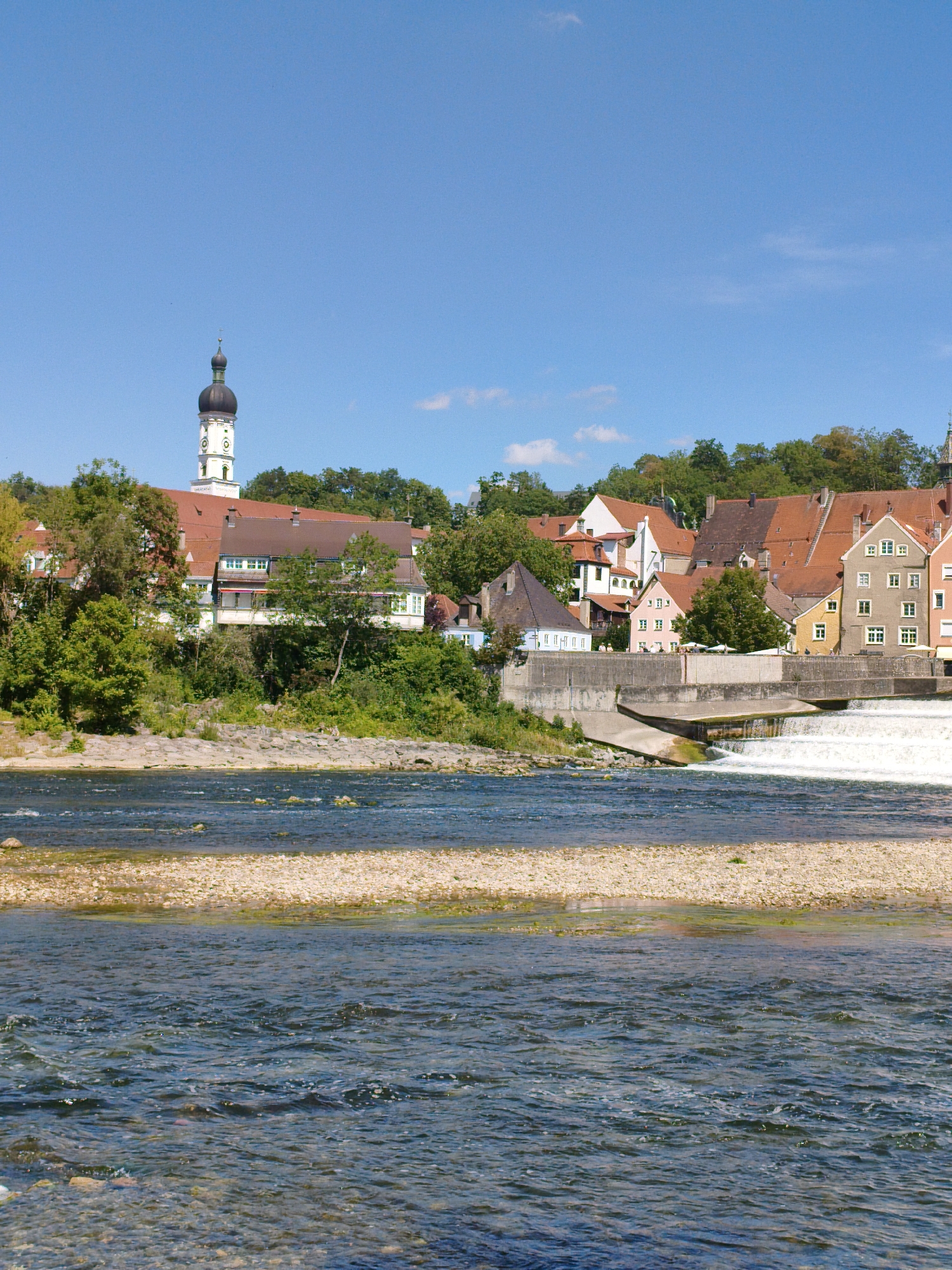 The image shows the Lech river in Landsberg am Lech, Germany. In the background, a town with traditional German architecture is visible, with buildings featuring red-tiled roofs and white facades. A prominent church spire with a black dome stands out against the blue sky. The river flows in the foreground, with a gravelly bank on the right side. A small waterfall or dam is visible in the middle ground, creating white water. Trees and greenery surround the town and line the riverbanks. The sky is clear and blue with a few wispy clouds. There are no people visible in the image.