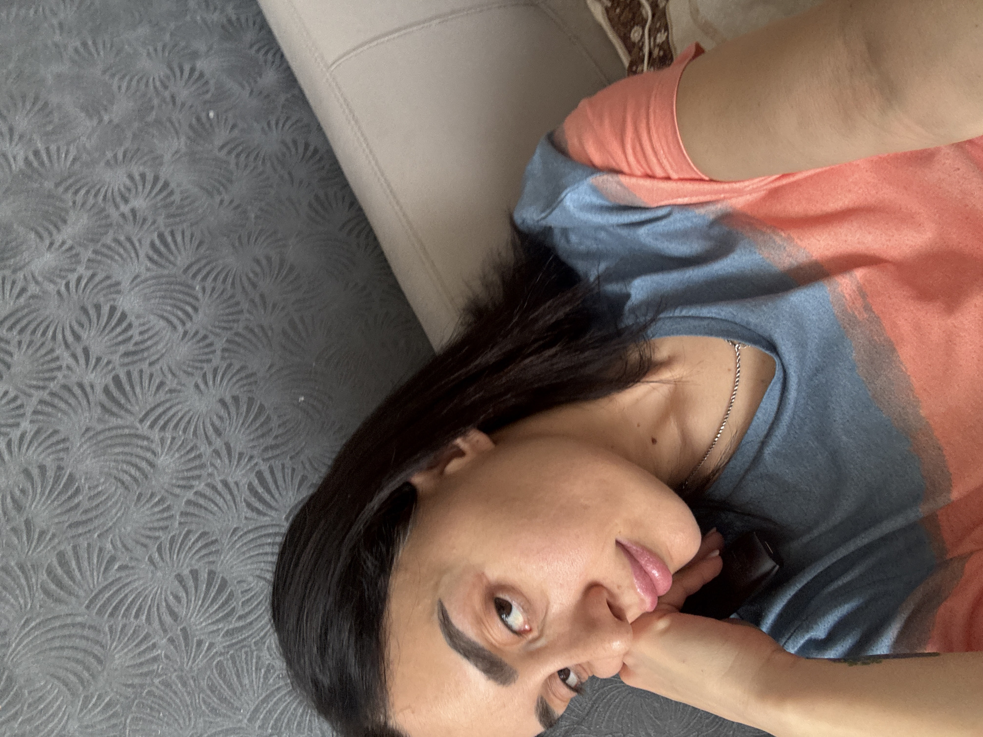 A woman with dark hair is lying on a patterned gray surface next to a beige upholstered object, possibly a sofa. She is wearing a salmon and blue t-shirt and a silver necklace. The location cannot be determined from the image alone.