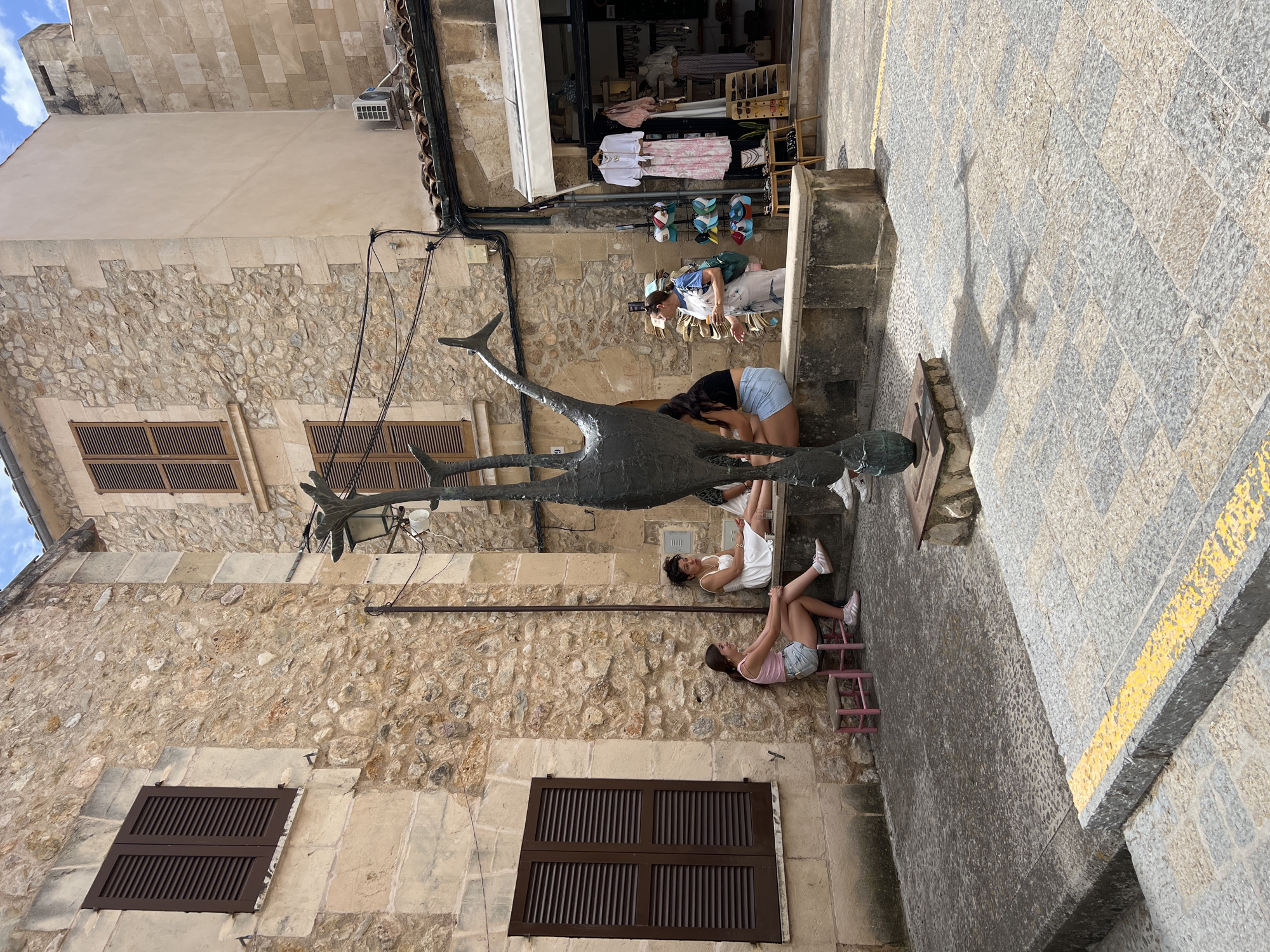 In the town square of Pollença, Spain, a group of people are seated on a stone bench next to a contemporary sculpture of an abstract bird. The sculpture is dark bronze, with elongated limbs and a slender body, casting a distinct shadow on the cobblestone pavement. To the left, two young women sit on small pink stools near a window with brown shutters. One woman wears a pink tank top and denim shorts, while the other has on a white dress. Across from them, further down the bench, another woman in denim shorts and a black top is seated. On the right side of the square, a woman wearing overalls stands near a shop that sells clothing and accessories. She is holding a drink in her hand. The buildings surrounding the square are made of rough-hewn stone, with wooden shutters on the windows. The ground is paved with irregular cobblestones. The sky is a clear blue with some light clouds.