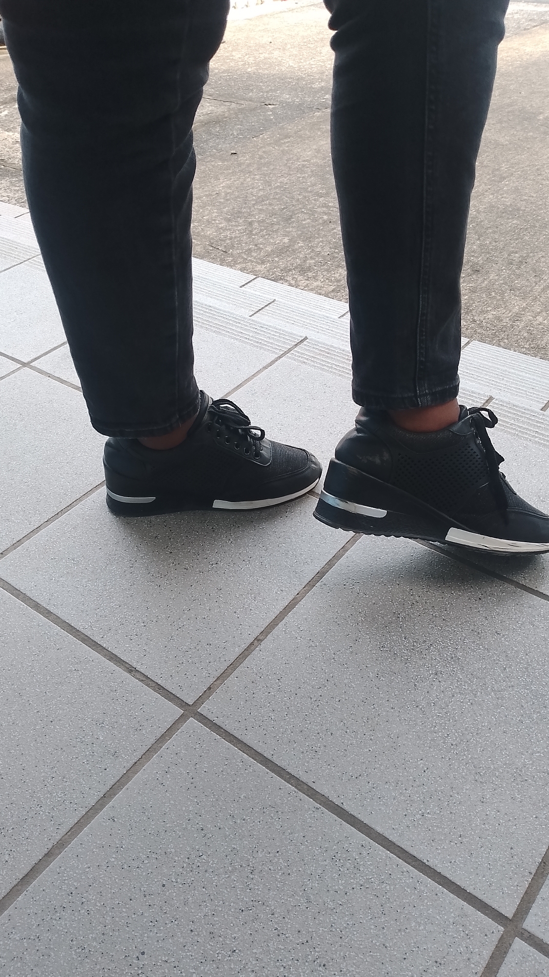 The image shows two feet wearing black sneakers with black jeans on a light gray tiled surface. The location is Orerokpe, Nigeria.