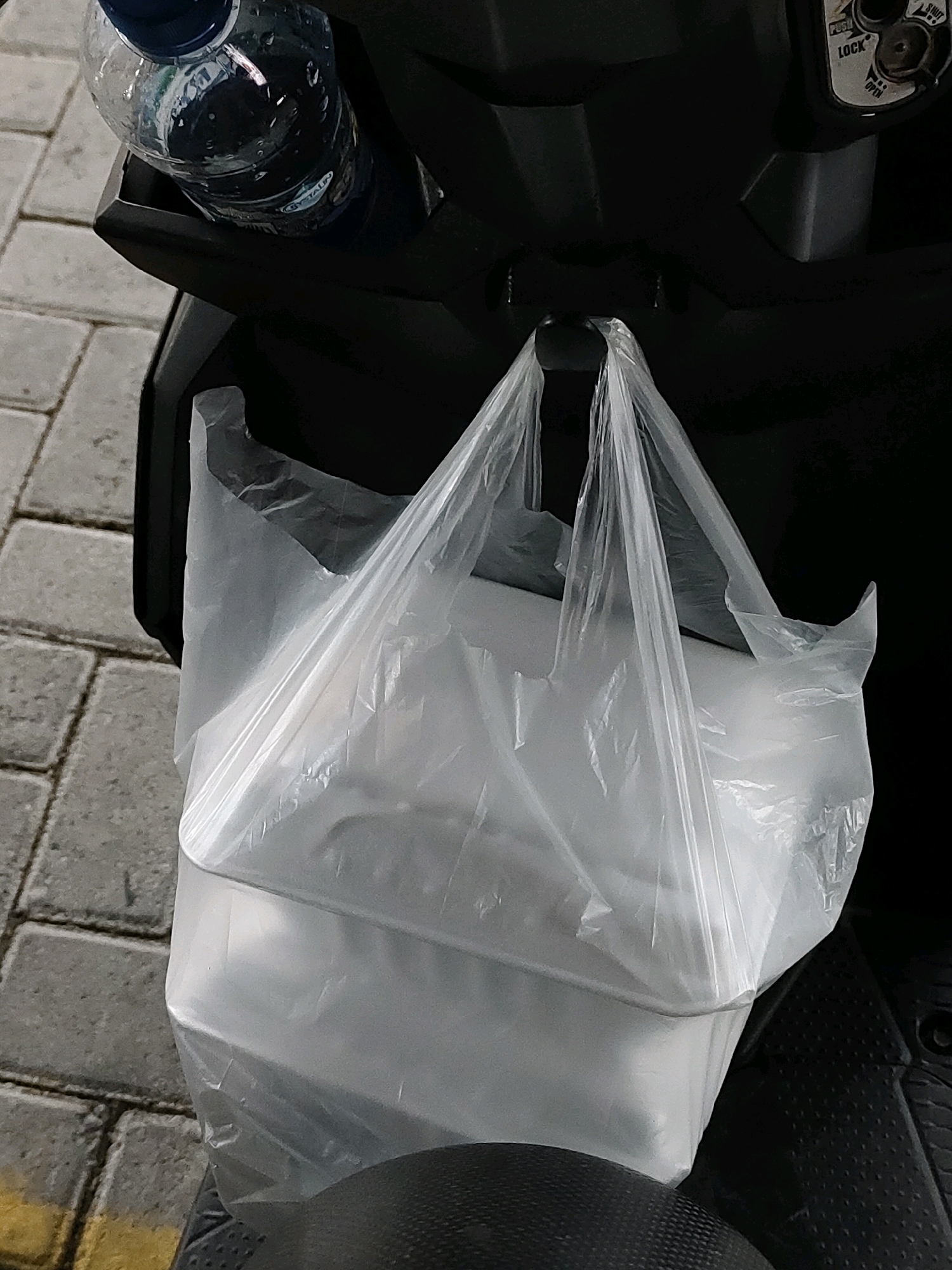 A clear plastic bag filled with white rectangular objects, likely food containers, hangs from a hook on the dashboard of a dark-colored scooter. A bottle of clear liquid with a blue cap rests in a compartment above the bag. The scooter is parked on a paved area with gray rectangular bricks, a portion of a yellow painted line is visible on the left. The background is dark, suggesting it is a vehicle's interior or a shaded area. The image appears to have been taken in Probolinggo, Indonesia, based on the context provided.