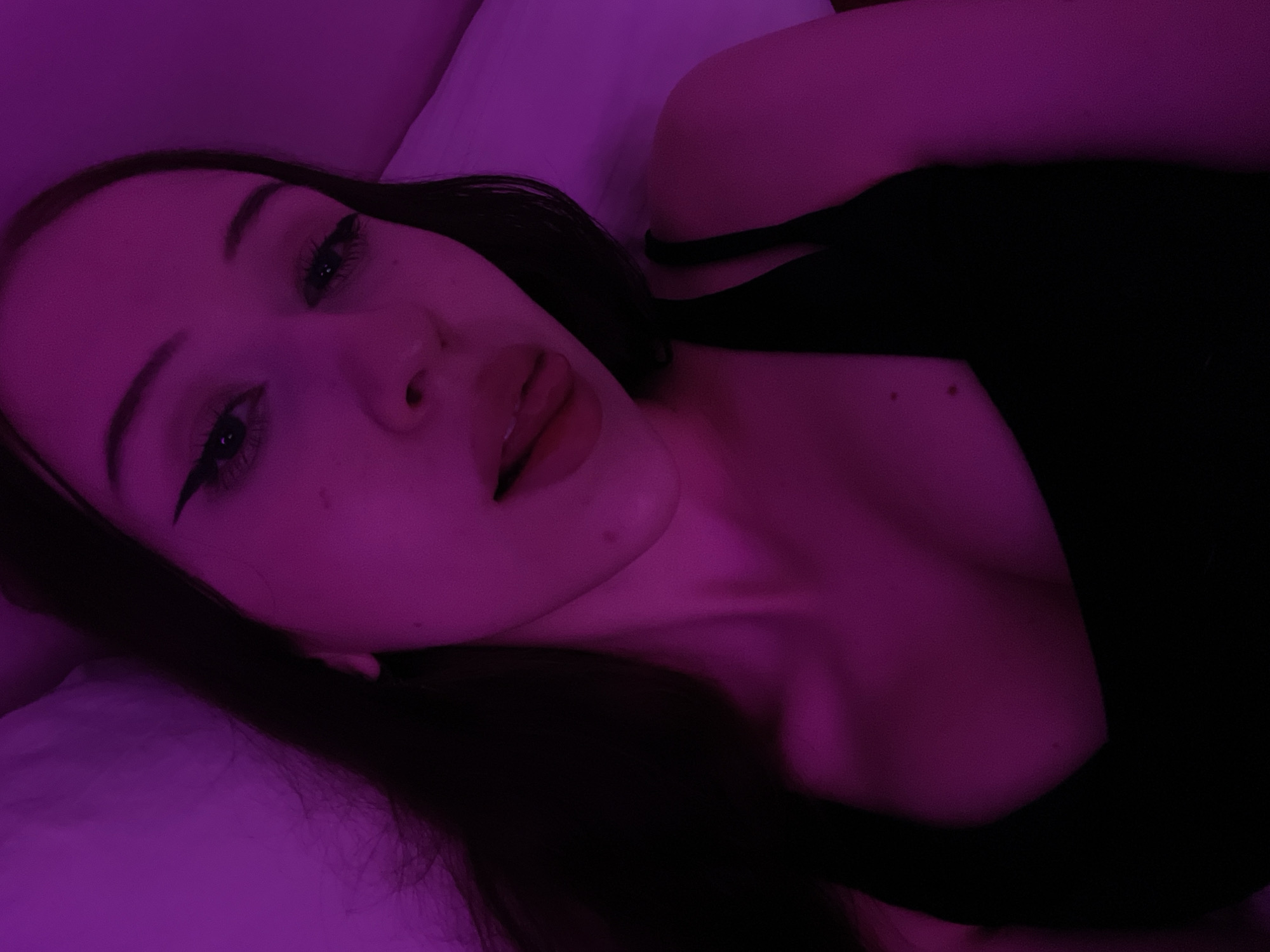 A young woman with dark hair, wearing a black tank top, is lying down. Her face is illuminated by purple light, and she is sticking her tongue out slightly. The background is a blurred pink and purple hue, suggesting an indoor setting. There is no information available about the location in Votkinsk, Russia.