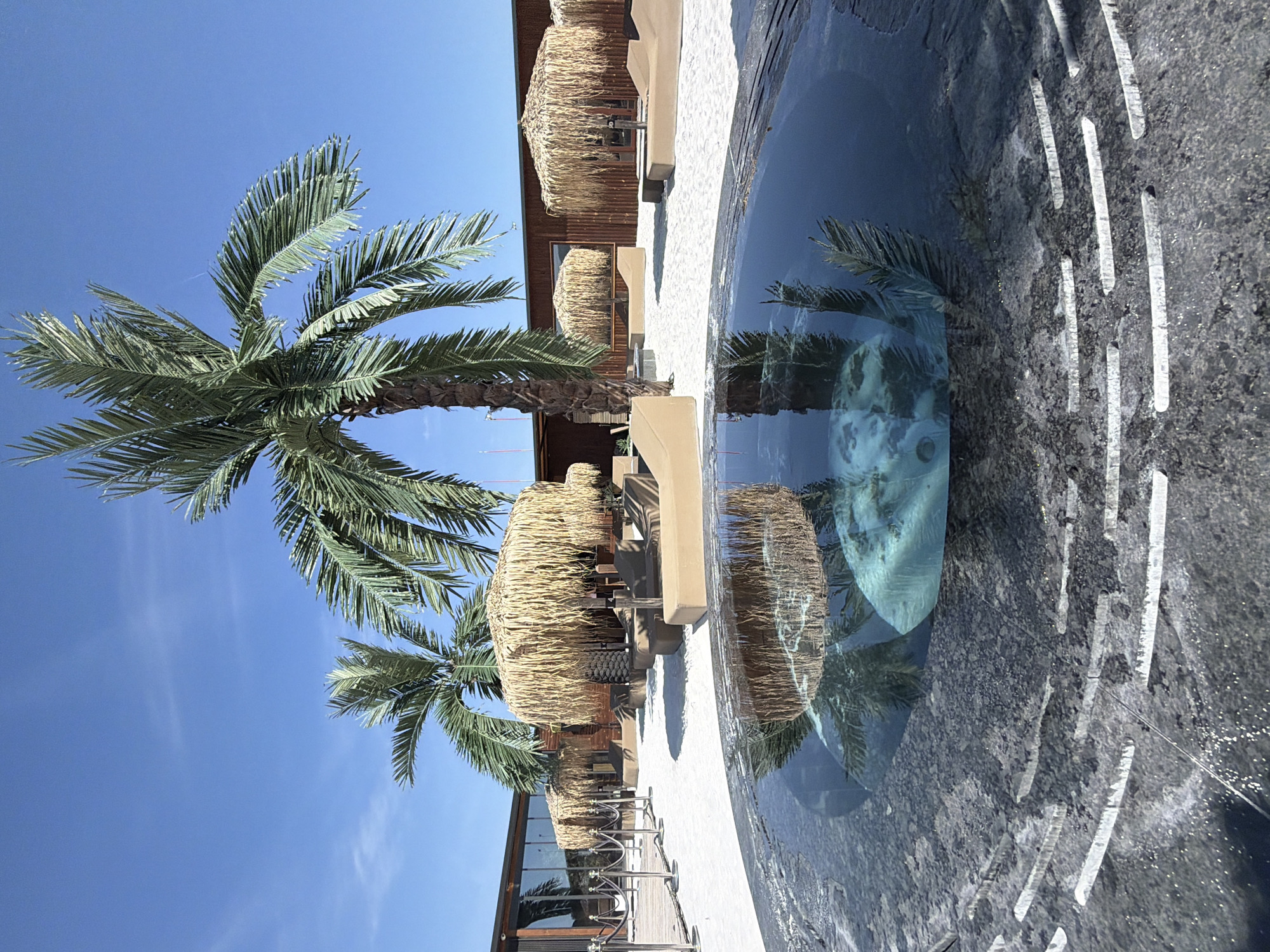 A circular water feature with a dark granite surround is in the foreground of this outdoor scene. The water is dark and reflective, showing an upside-down image of palm trees, thatched umbrellas, and loungers. The background features a sandy area with several thatched umbrellas providing shade over beige loungers. Palm trees are scattered throughout the scene. A modern building with large glass windows and a dark wooden exterior is visible behind the loungers and umbrellas. The sky is a clear, bright blue with a few wispy clouds. The location is identified as the city of Kazichene, Bulgaria.