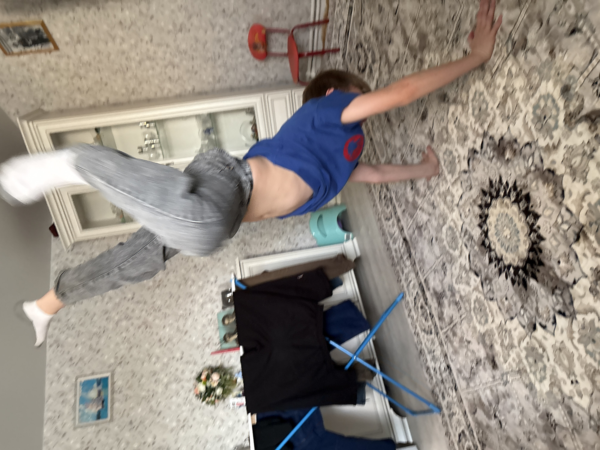 A person wearing a blue shirt and grey jeans is positioned mid-air, oriented horizontally. They are wearing white socks. Their hands are touching a carpeted wall. The background includes a white display cabinet, a small red chair, a white dresser with clothes drying on a blue rack, and patterned wallpaper. A picture hangs on the wall. The location is identified as Chaikovsky, Russia.