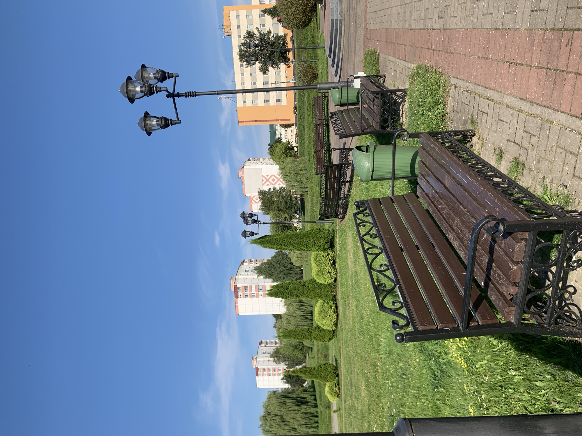 The image depicts a park scene in Slutsk, Belarus, under a clear blue sky with scattered clouds. In the foreground, a dark brown wooden bench with ornate black metal detailing sits on a brick pathway. To the right of this bench, another similar bench is visible, along with a green trash receptacle. Further back, a multi-headed black lamppost stands tall, casting a shadow. Beyond the pathway and benches, a lush green lawn is dotted with small yellow flowers. A line of neatly trimmed evergreen trees and rounded bushes forms a decorative border. In the mid-ground, more trees, including weeping willows, are present. The background features several multi-story apartment buildings with white and orange facades, characteristic of Soviet-era architecture. One building has a distinctive geometric pattern on its side. The overall impression is one of a well-maintained urban park on a sunny day.