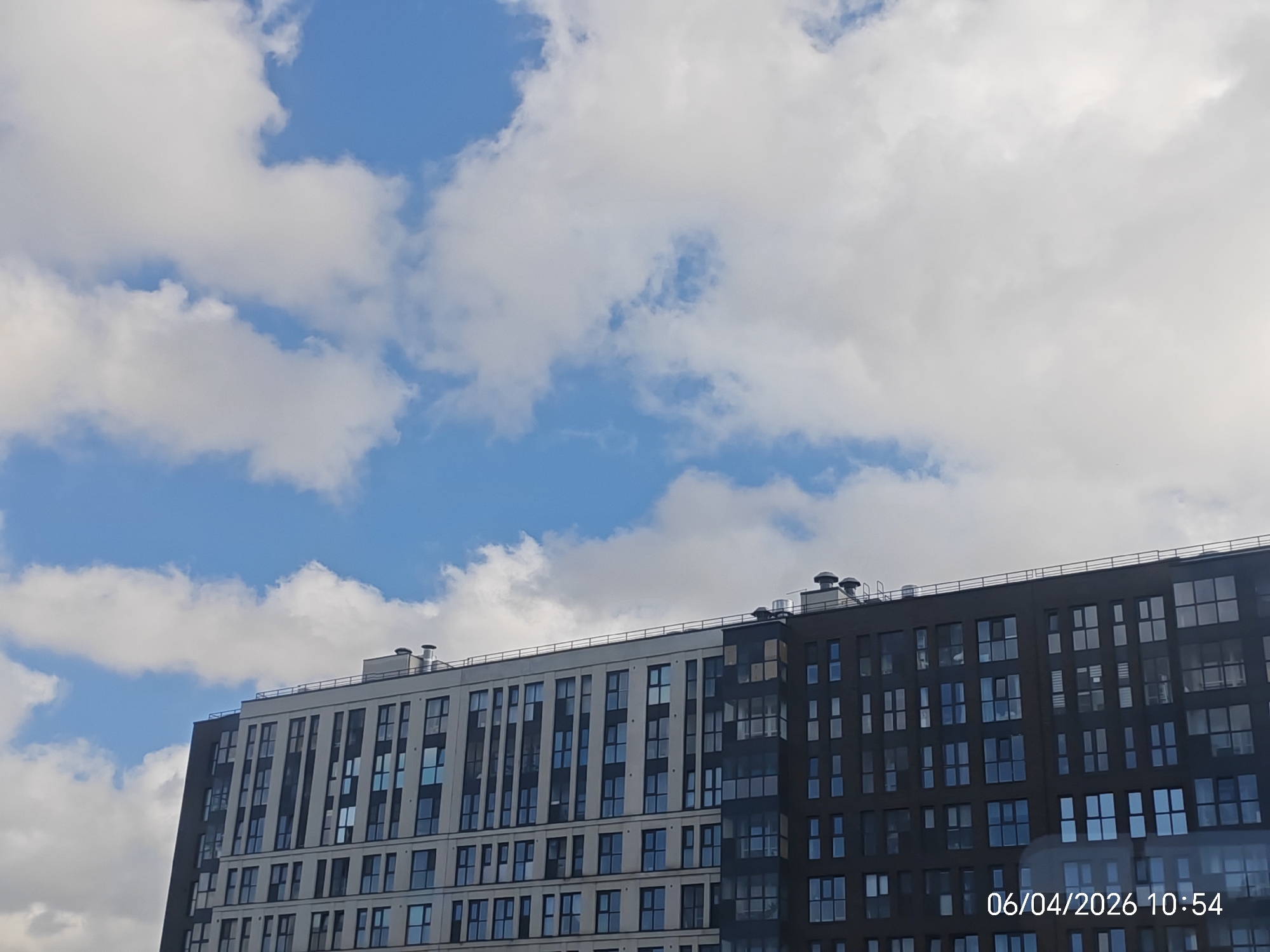 The image depicts a modern, multi-story apartment building against a backdrop of a partly cloudy blue sky. The building is divided into two distinct sections, one section with a lighter facade and the other with a darker facade. The windows are regularly spaced across both sections. The image also contains a timestamp "06/04/2026 10:54" in the lower right corner. The location of the image is Unknown, Unknown. There are no people visible in the image.