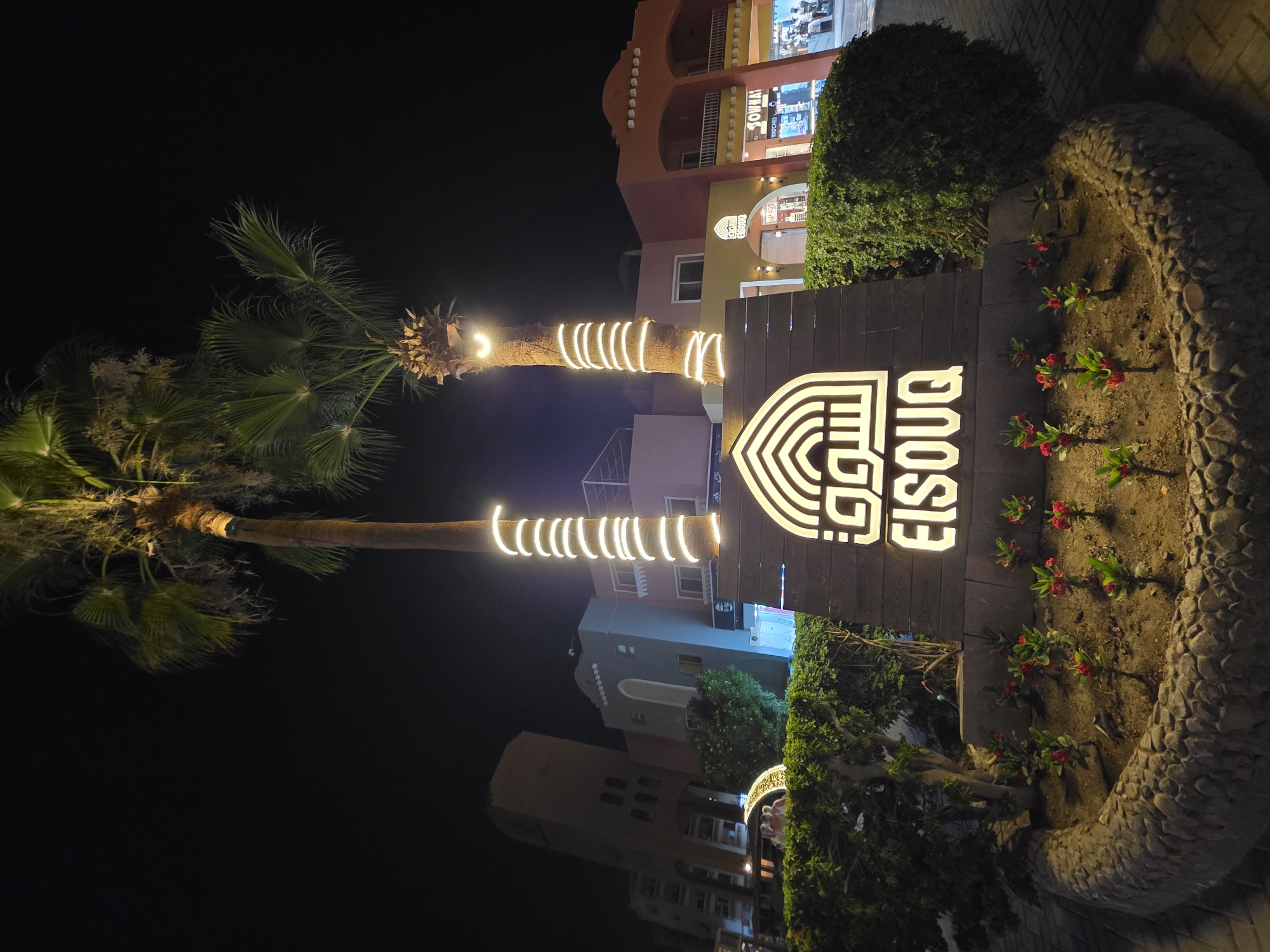 This nighttime outdoor shot shows a lit sign for "El Souk" in Hurghada, Egypt. The sign is placed in a garden bed with small flowering plants and a stone border. Behind the sign are palm trees wrapped with decorative lights. In the background are buildings, including a multi-story structure with shops. The overall environment suggests a tourist or commercial area at night.
