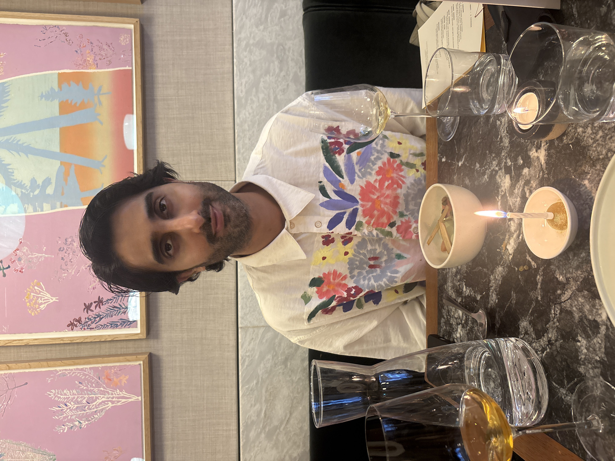 The image depicts a man seated at a marble table in what appears to be a restaurant. He is wearing a white button-down shirt adorned with a colorful floral print. In front of him, several glasses are visible, including a wine glass with a pale yellow liquid and a glass carafe filled with water. A small bowl with what looks like food and chopsticks sits on the table, along with two lit candles in small holders, one of which has a single candle on a stick. A menu or piece of paper is also present on the table. Behind the man, two framed artworks with botanical and abstract designs are hung on a textured wall. The overall setting suggests a dining experience, possibly a celebration given the candles. The man is looking directly at the camera with a slight smile.
