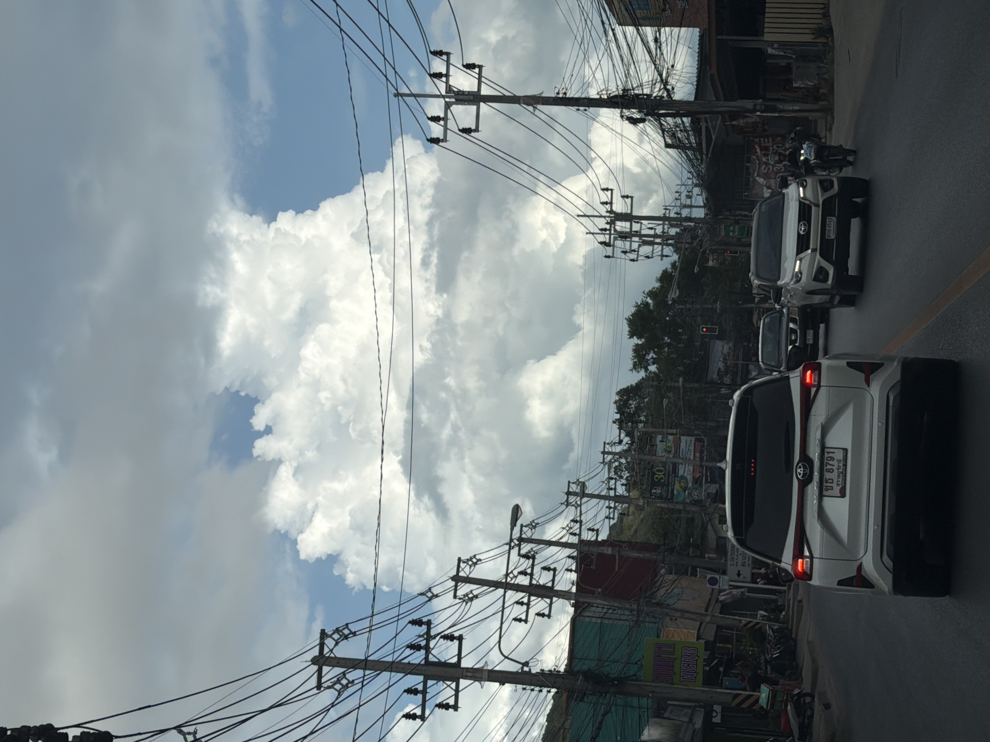 The image captures a street scene in the Ko Samui District of Thailand, under a partly cloudy sky. A white Toyota Veloz is in the foreground, facing away from the camera. To its right, a white Toyota Fortuner is visible, heading in the same direction, with a motorcycle and rider to its right. Several utility poles with numerous wires are strung along both sides of the street, suggesting a densely populated urban or semi-urban area. Buildings line the right side of the road, some appearing to be shops with signage. Trees are also present, indicating a tropical environment. The road surface is asphalt, with lane markings visible. The overall impression is of a typical street scene in a bustling town, possibly during the day, given the lighting.