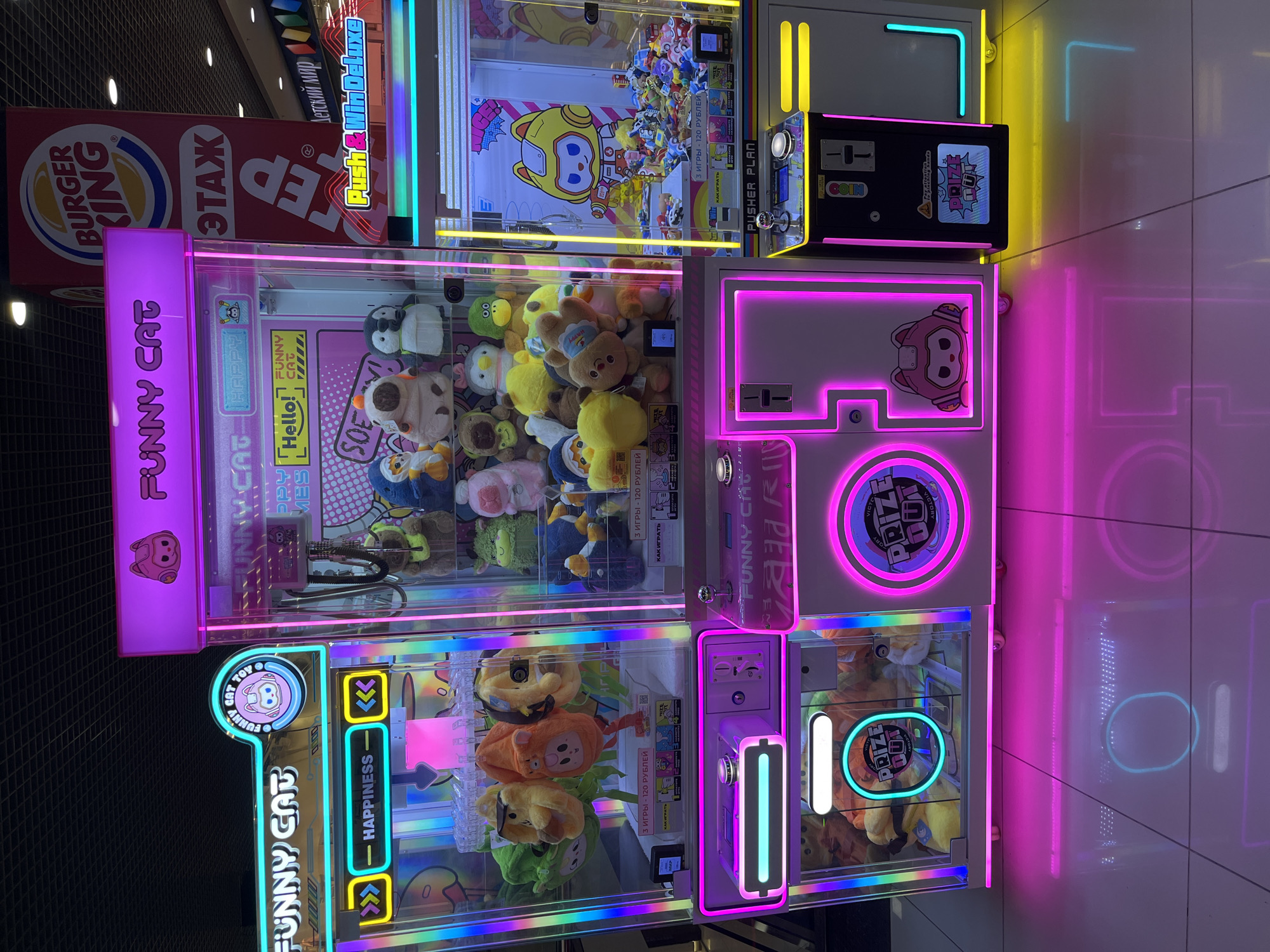 The image shows three "Funny Cat" crane machines stacked together in a brightly lit indoor location. The machines are illuminated with neon lights in pink, blue, yellow and green. Each machine contains plush toys, including cats, ducks, penguins and bears. Text on the machines, written in both English and Russian, provides instructions and pricing information. Above the crane machines are logos for Burger King and Push & Win Deluxe. Based on the Cyrillic text present, it is likely this image was taken in a Russian-speaking region, specifically the text implies the city of Izhevsk, Russia. The floor is made of light gray tiles.