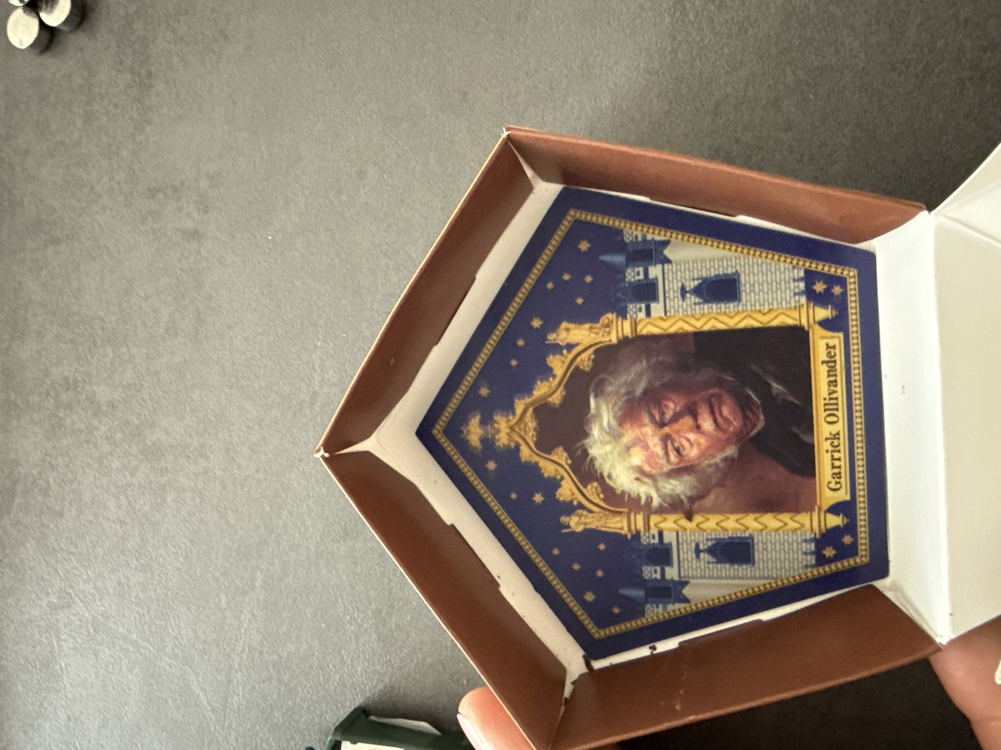 The image shows an open chocolate frog card inside its brown box. The card features Garrick Ollivander. The background is a dark gray surface. A few small, round objects are in the upper left corner. In the lower left corner, partially obscured, is a green object. The scene is indoors with artificial lighting. There is no indication of the city of Krasnoyarsk, Russia.