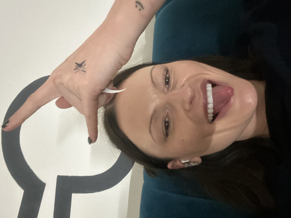 A close-up, selfie-style indoor photograph features a female subject with dark hair, fair-to-medium complexion, making a playful expression by sticking out her tongue. Her left ear, equipped with a white wireless earbud and a metallic ear cuff, is partially visible. Her right hand is raised above her head, forming a "rock on" gesture with extended thumb, index, and pinky fingers. Her fingernails are painted dark. Tattoos include a stylized starburst on the back of her right hand and a small smiley face on her right forearm/wrist. The subject is seated against a teal upholstered background, with a partial, large dark grey "G" logo visible on a white wall behind her. This logo design is consistent with branding found in a Google office, placing the location in the city of London, United Kingdom.
