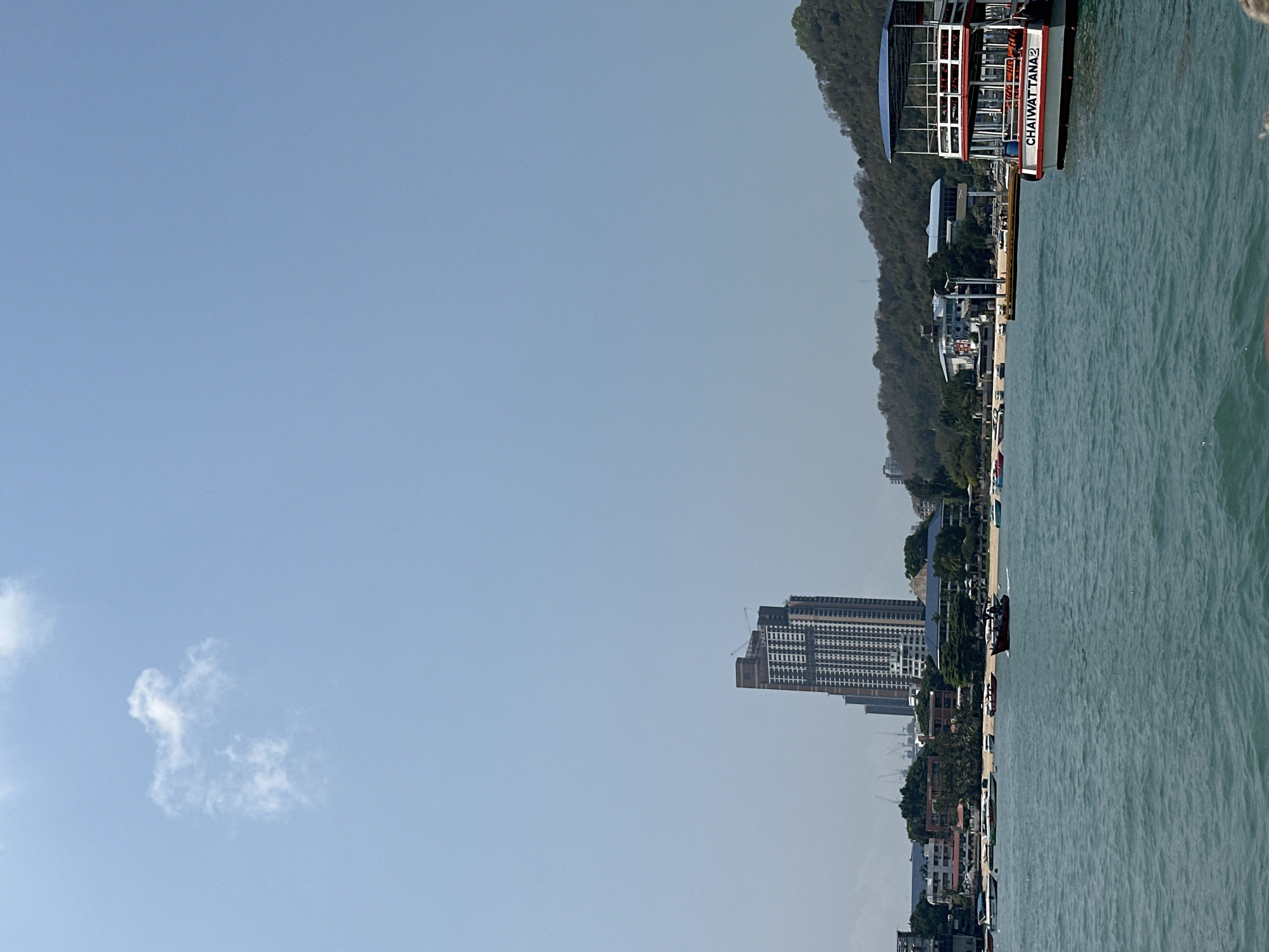 This image captures a coastal view in Pattaya City, Thailand. The foreground displays the ocean's surface, transitioning towards the shoreline with buildings, trees, and some vegetation. A high-rise building is visible amidst the city's architecture. In the upper-right corner, part of a boat can be seen along the coastline. The backdrop consists of a clear sky with a small cloud formation.