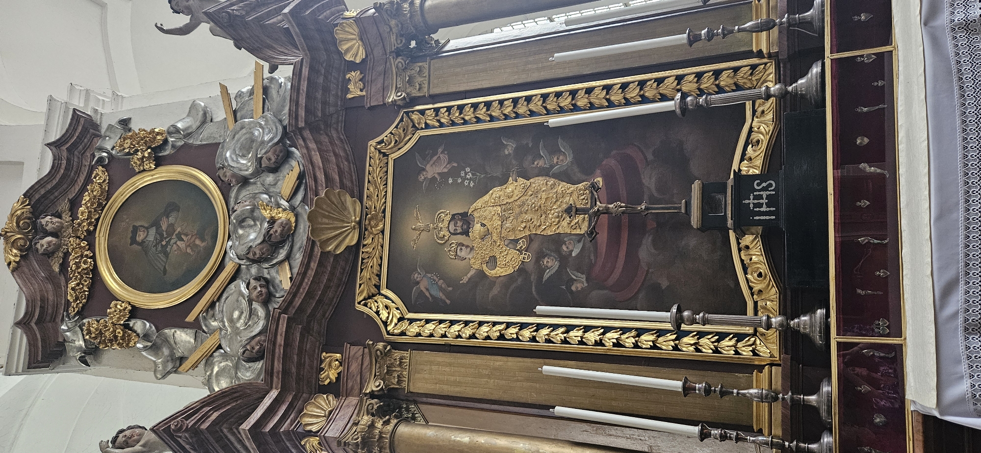 The image shows a close-up of an ornate altar or shrine, likely within a church in Hrodna, Belarus. The main focus is a large, framed painting depicting religious figures. The painting is richly detailed, featuring a central figure adorned in a golden, elaborately patterned robe and wearing a crown. This figure holds a crucifix and a staff. Above the crowned figure is a dove, representing the Holy Spirit. Surrounding the central figures are several cherubic angels with small wings. The painting is set against a dark, cloudy background, evoking a celestial or heavenly scene.
To the left of the main painting, a smaller circular portrait is visible, framed in gold. This portrait shows a depiction of a mother and child, also rendered in a painterly style.
The altar itself is highly decorative, with gilded carvings and sculpted elements that appear to represent cherubs or angels. The color palette of the altar is dominated by deep browns, gold, and silver. Several large white candles in ornate silver holders are positioned in front of the painting, suggesting the altar is in active use.
To the right of the main painting, a dark, rectangular structure is visible, featuring the Latin abbreviation "IHS" in white lettering. Adjacent to this is a display case with a velvet-lined interior, containing small, silver reliquaries, possibly representing body parts or symbols of saints. These are arranged in rows, adding to the sacred nature of the display. A white cloth with lace trim hangs on the far right edge of the image. The lighting suggests an interior setting, likely during the day, but without any indication of natural light. The overall impression is one of a richly adorned and sacred space, likely a significant religious site.
