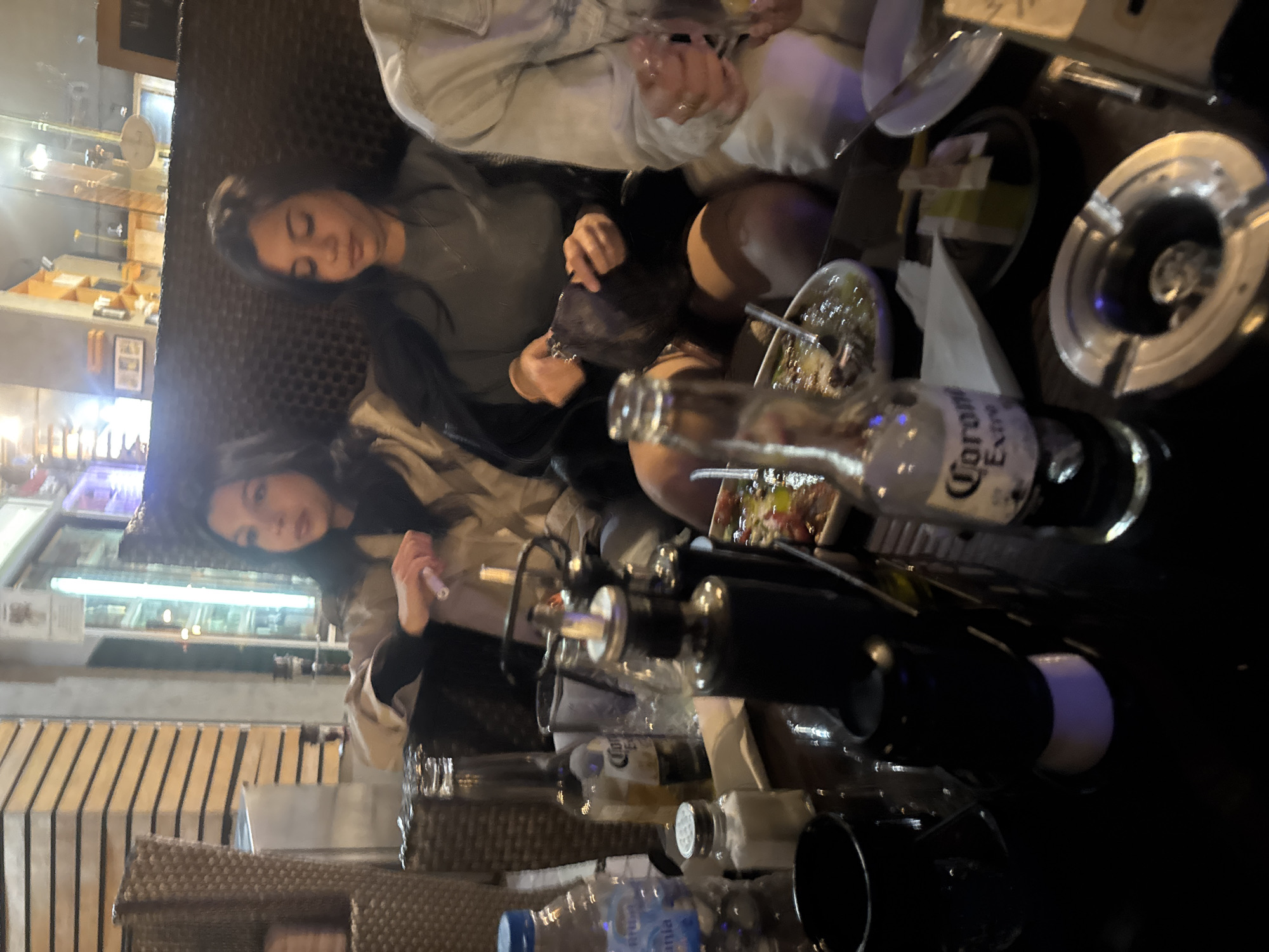 The image depicts an indoor setting, possibly a restaurant or bar, with two women seated at a table. One woman wears a light-colored jacket and a dark top, while the other wears a grey top and a black jacket. Various objects are visible on the table, including a Corona Extra beer bottle, a silver ashtray, plates, and what appears to be food in a bowl. Other unidentified bottles and glassware are present. In the background, through a window or opening, a brightly lit display with shelves and various items is seen. The scene appears casual, with individuals interacting and items scattered on the table. There is no explicit information in the image to determine if the location is in Kyustendil, Bulgaria.
