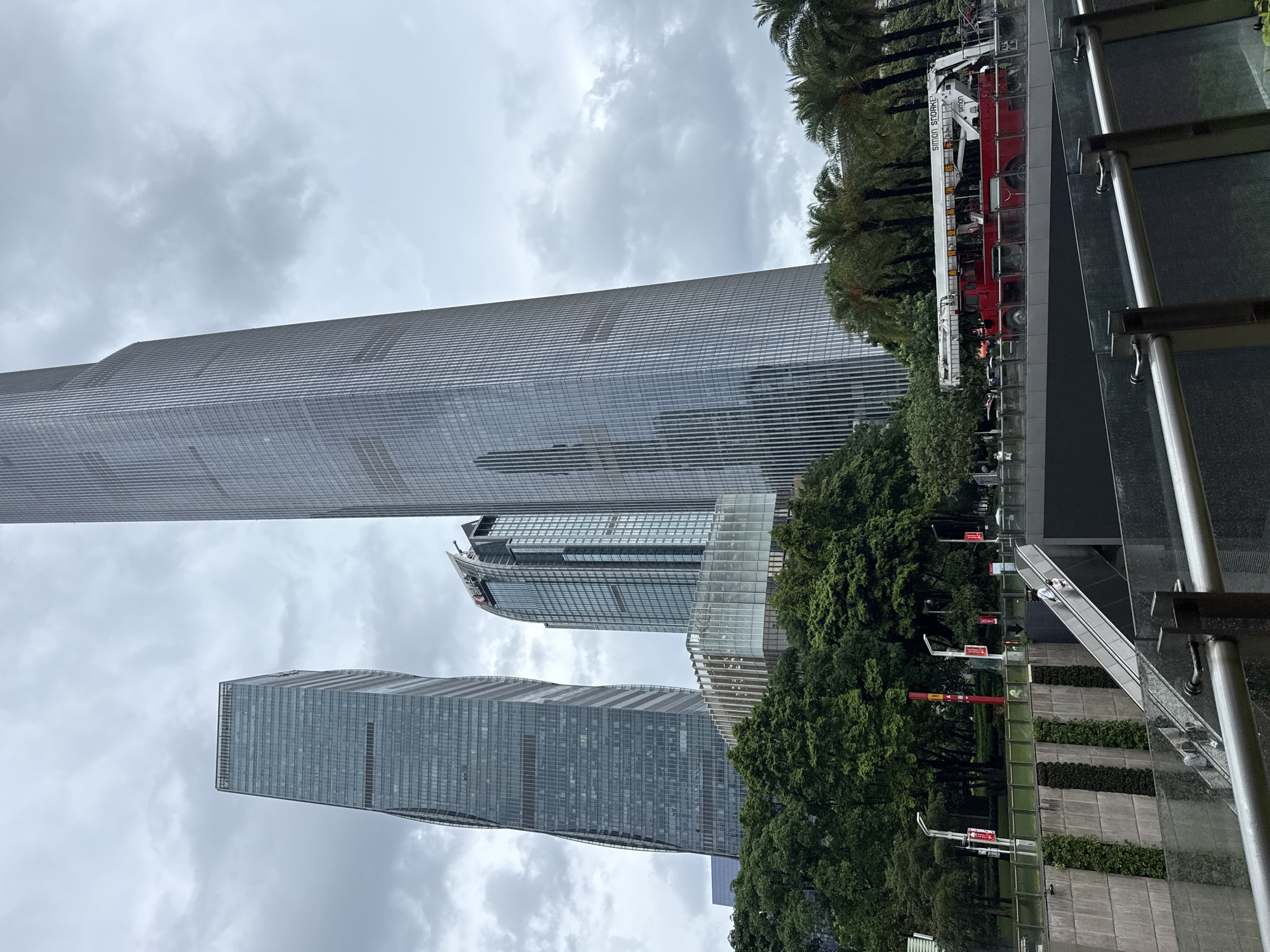 The image captures a view of modern skyscrapers in Tianhe District, China, under an overcast sky. In the foreground, a red fire truck with "SIMON SNORKEL" written on its side is parked on a bridge, partially obscured by a glass railing. Below the bridge, an outdoor escalator carries two individuals upwards, and another escalator is visible leading down into a darker area. To the left of the escalators, a concrete wall is adorned with vertical green plant installations. Behind the wall, lush green trees form a dense foliage line. In the background, three prominent skyscrapers dominate the skyline, with the tallest, a sleek glass building, in the center-right. To its left, a slightly shorter, similarly modern skyscraper stands, and further left, a distinct, twisted glass tower is visible. The overall scene depicts an urban environment characterized by contemporary architecture and greenery.