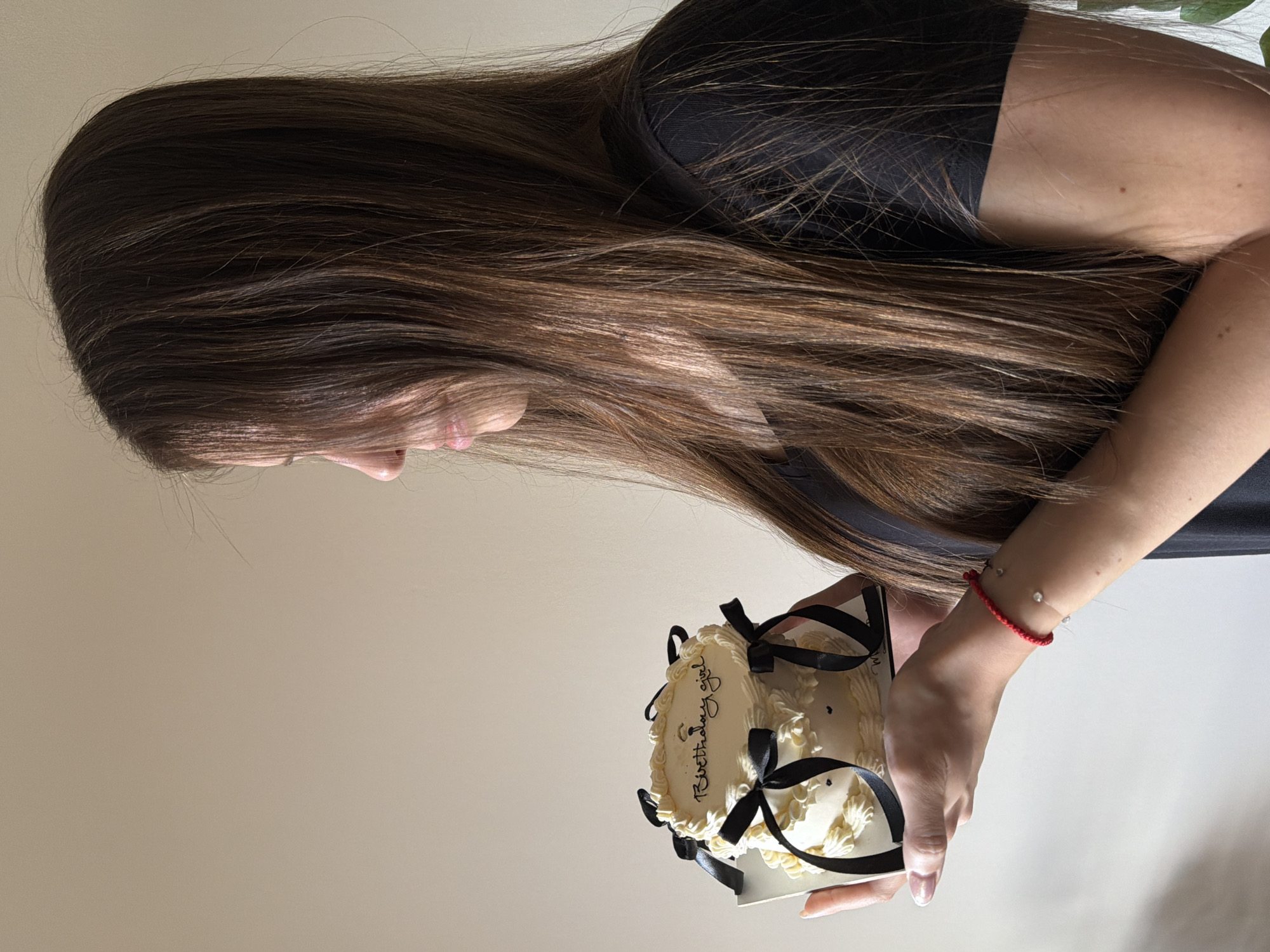 A young adult female, depicted in profile, occupies the right half of the frame, facing right and looking downwards. She possesses long, straight, dark brown hair with lighter brown highlights, which partially covers her right cheek and chin. She is wearing a dark, possibly black, short-sleeved garment. Her left hand and forearm are visible, supporting a small, two-tiered birthday cake resting on a silver-colored cake board. A red string bracelet adorned with two small, clear, faceted beads is visible on her left wrist. The cake is decorated with cream-colored frosting, piped in swirls and rosettes, and features multiple black satin ribbon bows around its tiers. The top tier of the cake has the phrase "Birthday girl" written in black script icing. The background is a plain, light beige wall, indicating an indoor setting with even lighting. The specific location in Sofia, Bulgaria, cannot be determined from the visual information provided in the image.