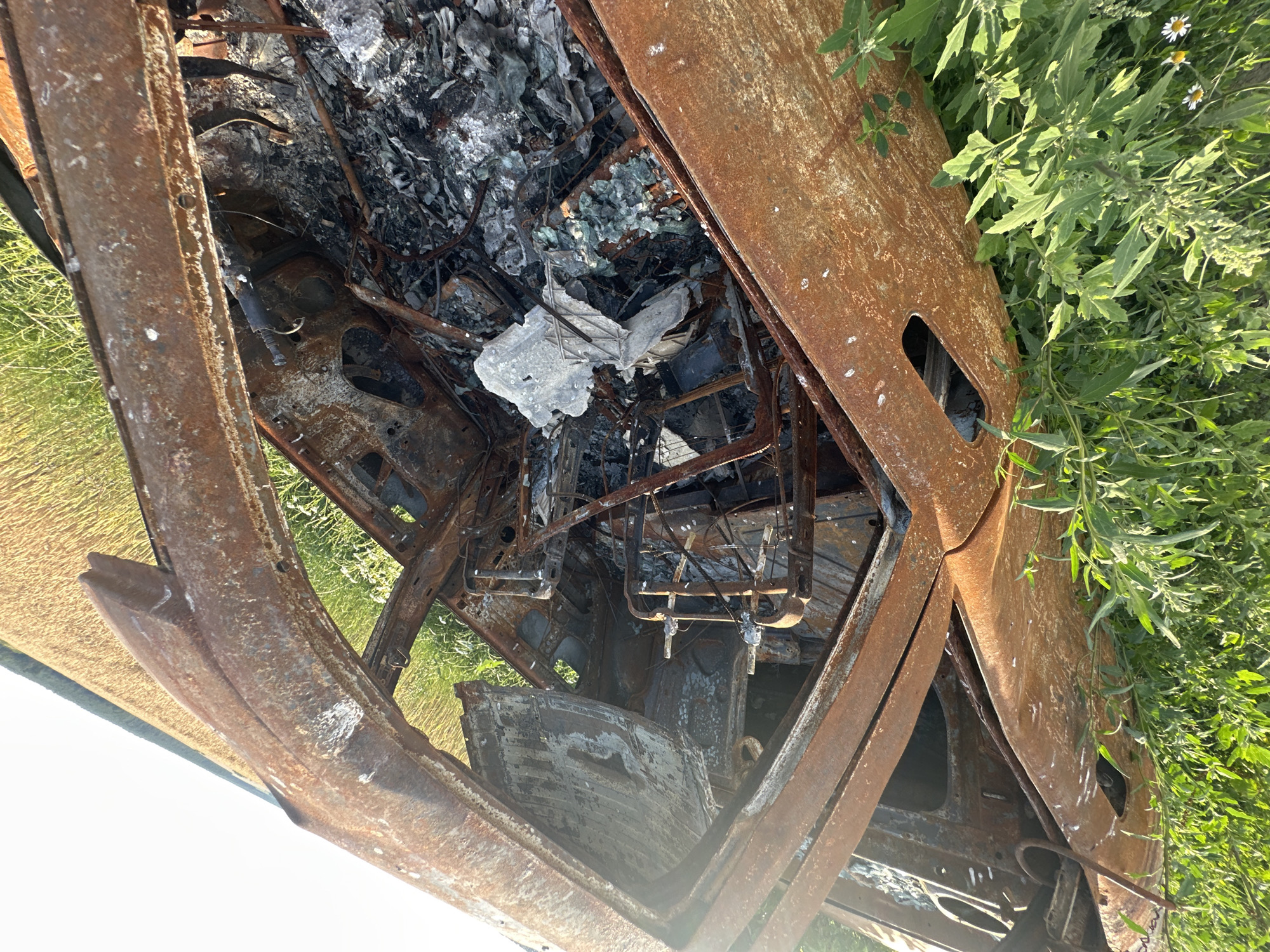 The image shows the interior of a severely damaged and rusted-out vehicle, likely a car, lying on its side in a grassy field. The vehicle's structure is heavily corroded, with exposed metal framing and evidence of significant heat damage, including melted or warped materials and ash-like debris filling the interior. The seats appear to be skeletal remains of metal.
The surrounding environment includes lush green vegetation and some small white flowers at the bottom of the frame. In the background, a field of golden crop, possibly wheat or barley, stretches towards a bright, clear sky. The scene is consistent with the outskirts of a rural area. The image does not depict any people or any specific identifiable landmarks within the city of Makiv, Ukraine, beyond the general context of a rural Ukrainian setting.