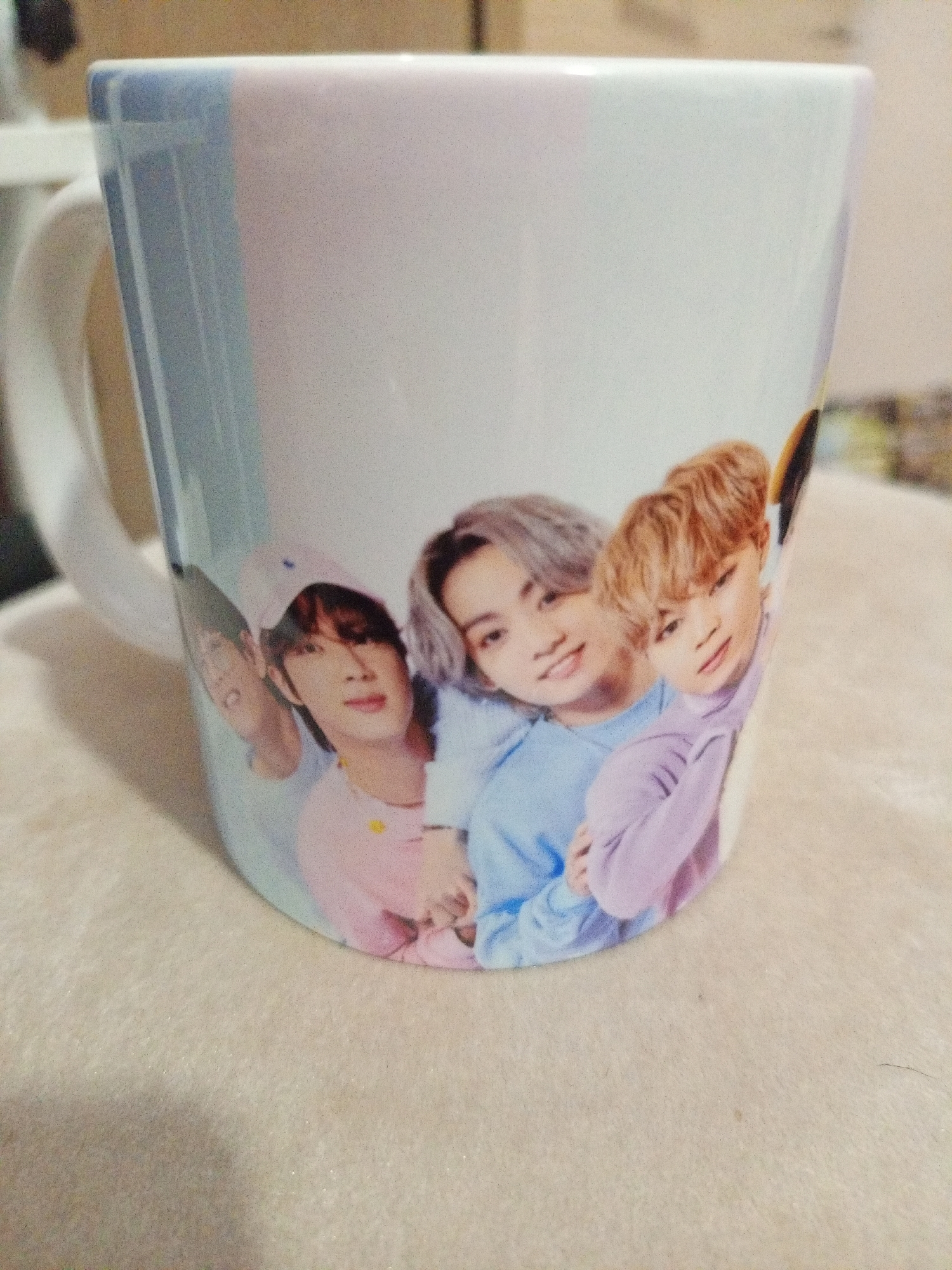 The image shows a white mug with a printed image of four young men. Three of the men are in focus, and one is partially visible on the left. The man on the far right has light brown hair and is wearing a light purple sweater. Next to him, to his left, is a man with graying hair wearing a light blue sweater. The man to his left has dark hair and is wearing a light pink shirt and a light pink baseball cap. The partially visible man on the far left appears to have dark hair. The mug has a light blue and white striped pattern at the top. The mug is placed on a textured, cream-colored surface, possibly a tablecloth or rug. The background is blurred and shows a hint of a white object on the left, possibly another mug or container, and a brown, out-of-focus background. The scene is indoors and well-lit. The image does not provide any information about the city of Otradnaya, Russia.