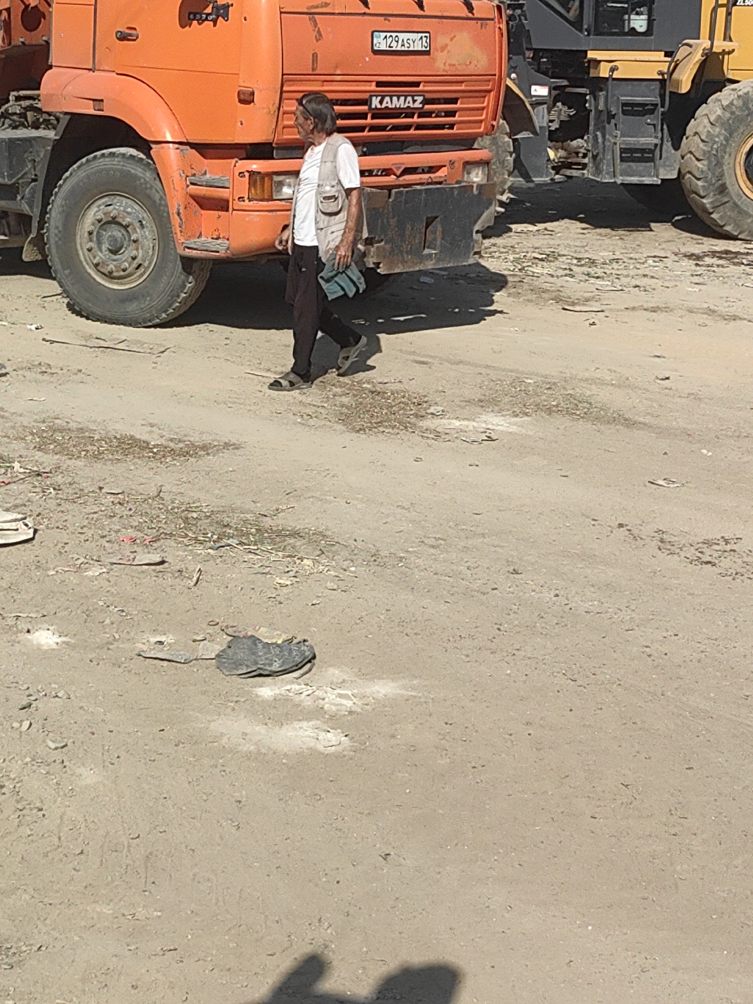 The image shows an elderly man walking in front of an orange KAMAZ truck and a yellow construction vehicle on a dirt lot in Shymkent, Kazakhstan. The man is wearing a white t-shirt, a light-colored vest, dark pants, and sandals. He is carrying a blue bag in his left hand. The truck has a license plate that reads "KZ 129 ASY 13". The ground is covered in dirt and scattered debris, including some flattened items that appear to be trash. A shadow of a person is visible in the lower right corner of the image. The scene appears to be an outdoor industrial or construction area.