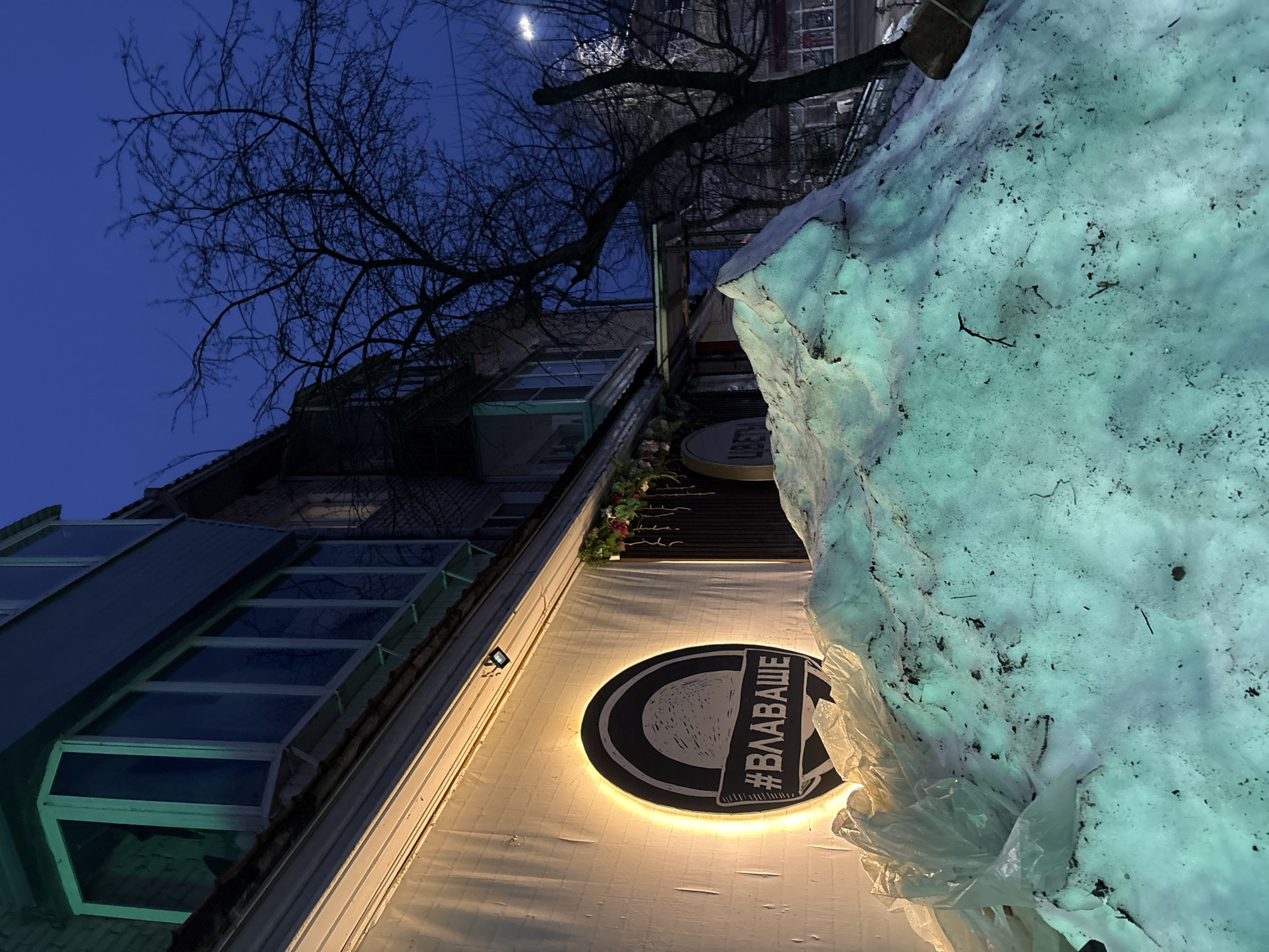 The image depicts a low-angle view of a building in Chaikovsky, Russia, at dusk or night. A large snowdrift is prominently featured in the foreground, partially obscuring the building's facade. The building's architecture is modern, with a sloped, glass-paneled roof section. A circular sign with Russian text is visible on the building's wall, illuminated from behind. A bare tree branch extends across the upper portion of the frame against the dark blue sky. The photograph appears to be taken from street level.