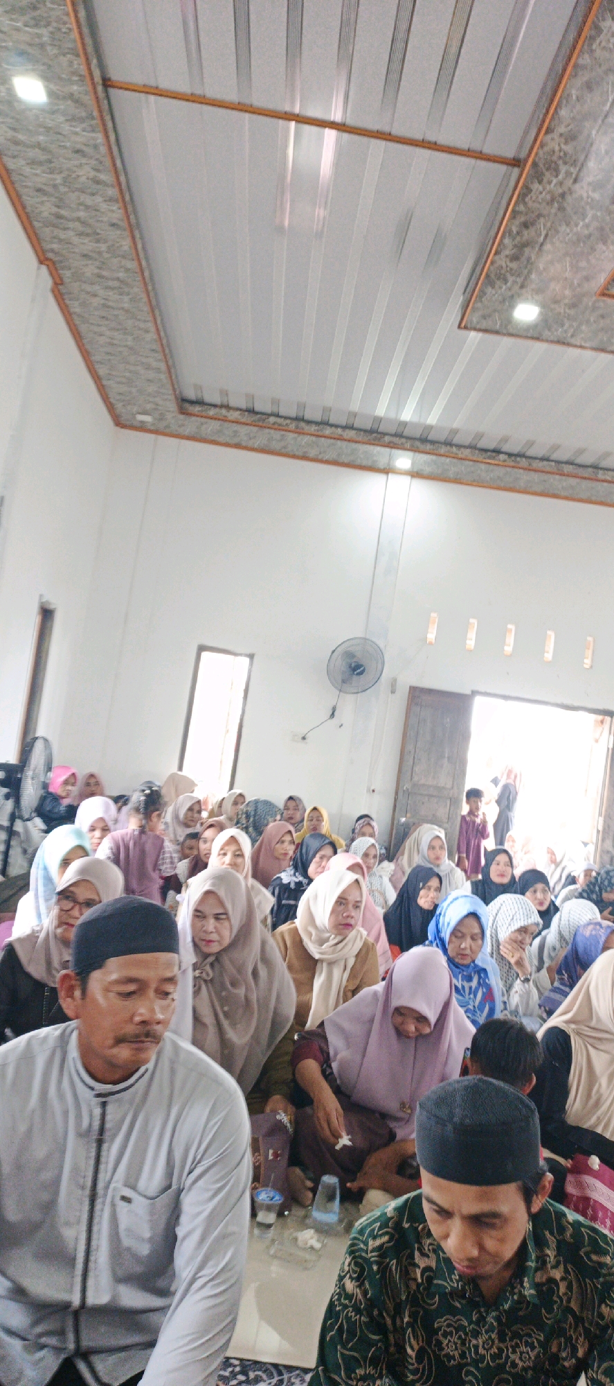 In Simbur Naik, Indonesia, a group of approximately 50 people, primarily women wearing hijabs, are seated on the floor of a room, attending an event. The room has white walls and a patterned ceiling with orange trim. In the foreground, a man in a grey collared shirt and a black cap is visible, along with another man in a green patterned shirt and black cap. Several women are seen in various colored hijabs, some looking towards the front of the room, others interacting with each other. A few children are also present within the gathering. The room appears to be a community space, possibly a mosque or a community hall, with windows and doors visible in the background. A ceiling fan is mounted on the wall. The lighting suggests it is daytime, with natural light entering from the windows and artificial light from overhead fixtures.