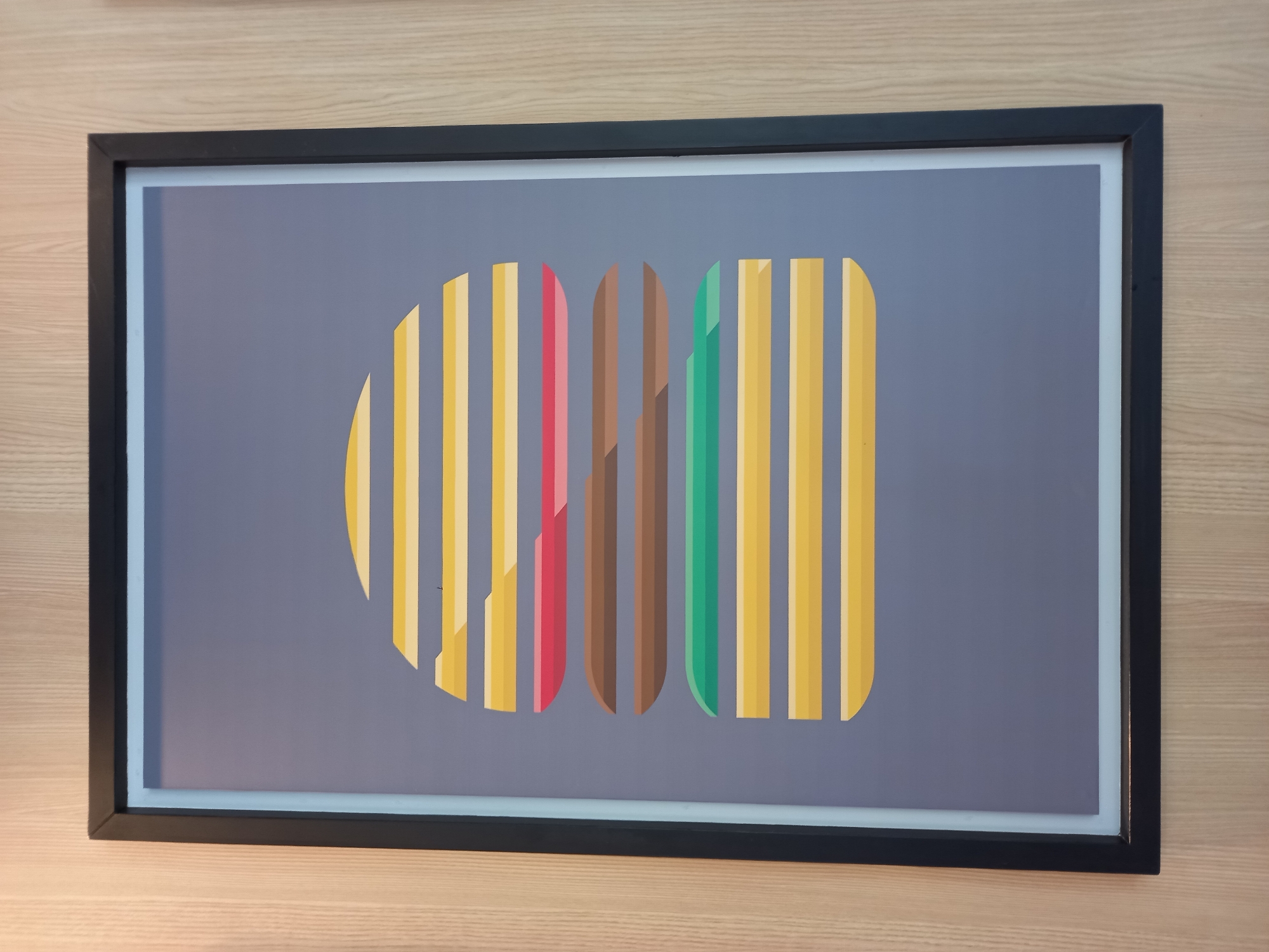 A framed artwork is hanging on a light wood-paneled wall. The artwork is a minimalist depiction of a hamburger. The background of the artwork is a muted gray. The hamburger is composed of horizontal stripes of various colors representing the different layers of a burger. Starting from the top, there are several yellow stripes, followed by a red stripe, then two brown stripes, a green stripe, and finally more yellow stripes at the bottom. The frame is black. The image is taken from a slightly downward angle. No people are visible. The location is the city of Baan Bo Phut, Thailand.