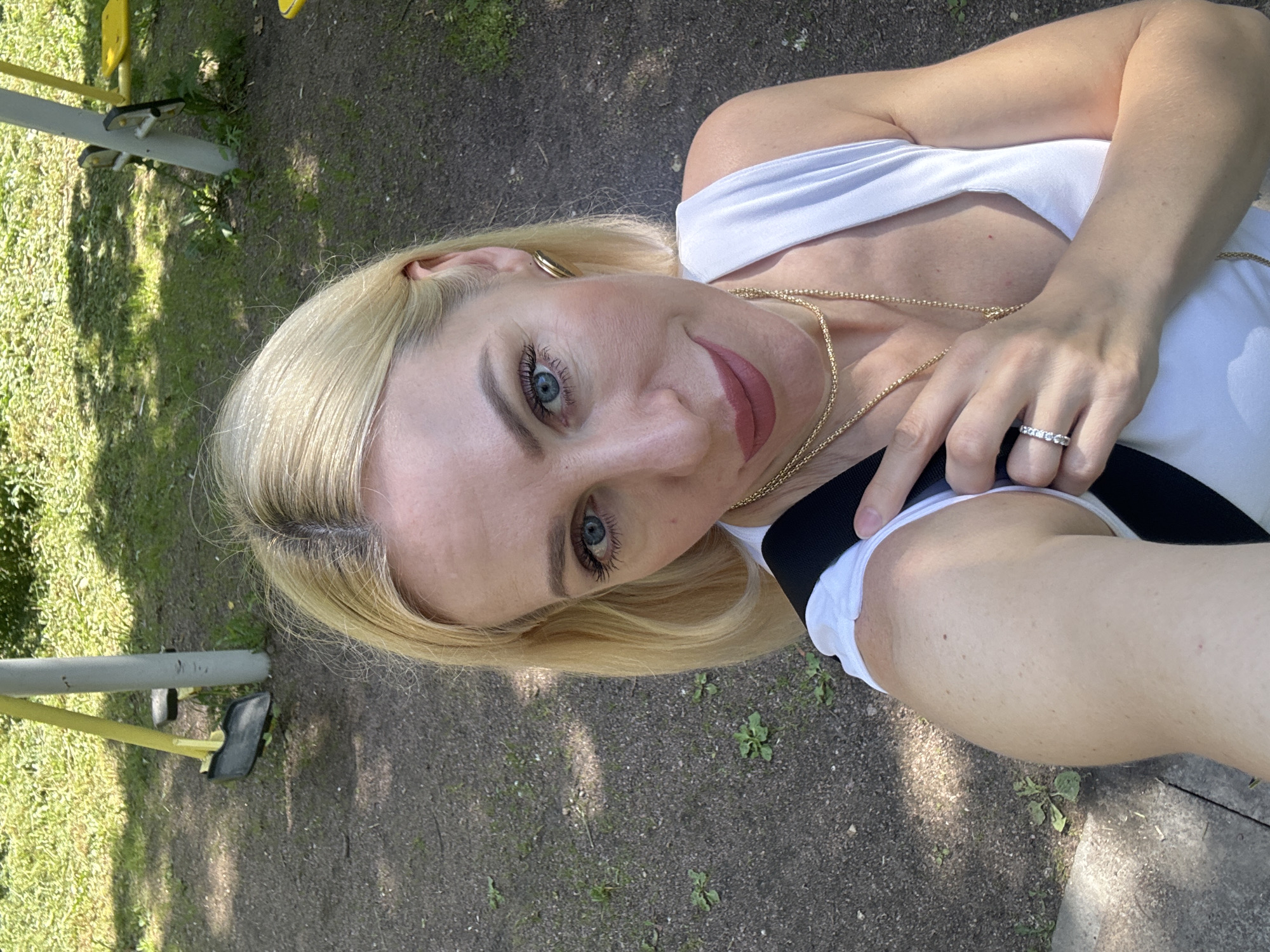 A blonde woman with blue eyes takes a selfie in an outdoor setting. She is wearing a white tank top with a black strap and gold necklaces. Her left hand is resting on her chest, displaying a diamond ring. The background appears to be a park or playground area with gravel ground, green grass, and playground equipment, including yellow swings and grey poles. The lighting suggests it is daytime.