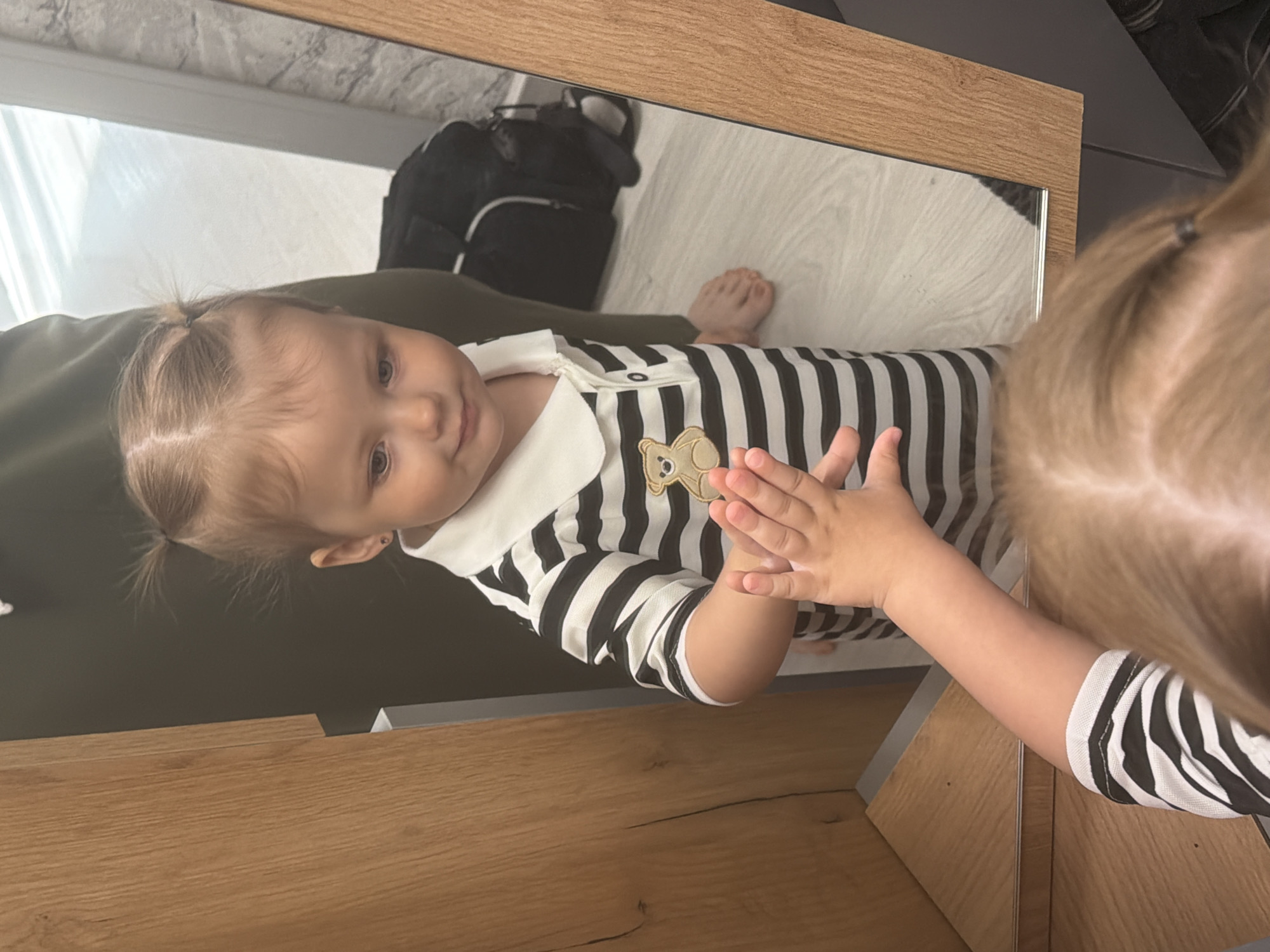 A baby girl wearing a black and white striped shirt with a white collar and a teddy bear patch is seen reflected in a framed mirror. The baby's reflection shows the girl touching her own reflection in the mirror. In the background of the reflection, there is a black backpack. The floor appears to be white or light-colored. The surrounding furniture or surfaces are wooden. The location is Reshetnikovo, Russia.