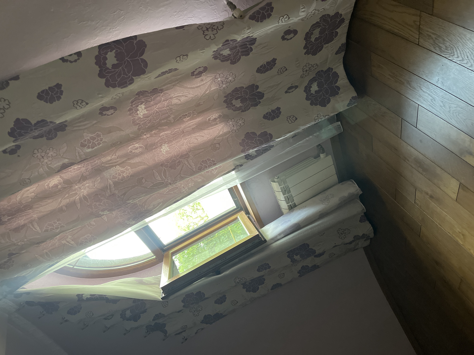 The image depicts an interior scene, likely a room, with a window that is partially open. Through the window, green foliage is visible, suggesting an outdoor environment with trees. The window has a wooden frame and is positioned at an angle, contributing to a slightly distorted perspective of the room.
Dominating the foreground and extending across the frame are curtains with a floral pattern, featuring large purple flowers against a lighter background. The curtains are partially drawn, with one side pulled back, revealing the window. The light streaming through the window illuminates the curtains and casts shadows on the wooden floor.
Below the window, a white radiator is visible. The walls of the room appear to be painted in a light, possibly pinkish, hue. The floor is made of wooden planks, laid horizontally and exhibiting a natural wood grain.
The overall impression is one of a softly lit interior, with the natural light from the window playing a significant role in the composition. The scene is static, with no people or explicit actions taking place. The environment appears to be residential, possibly a bedroom or living area.
The image does not provide any specific details or visual cues that would allow for the identification of its location within the city of Tarnivtsi, Ukraine.