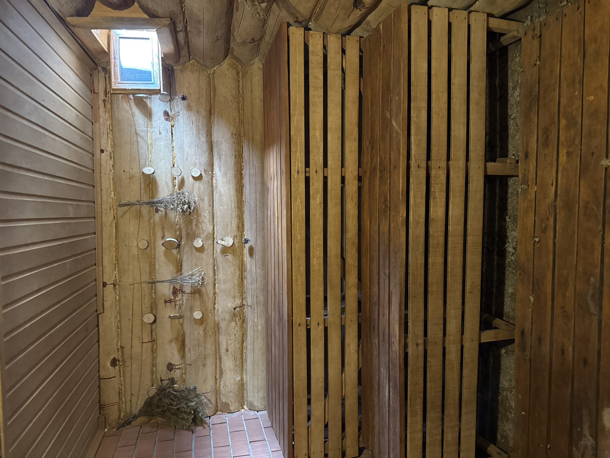 A photograph taken indoors in a room in Kordonnyi, Russia, shows a sauna. The walls are paneled with wooden planks. The room has a brick floor, and a small window in the upper left corner. On one wall are some dried herbs and wooden pegs. A partial privacy screen made of spaced wooden slats stands on the right.