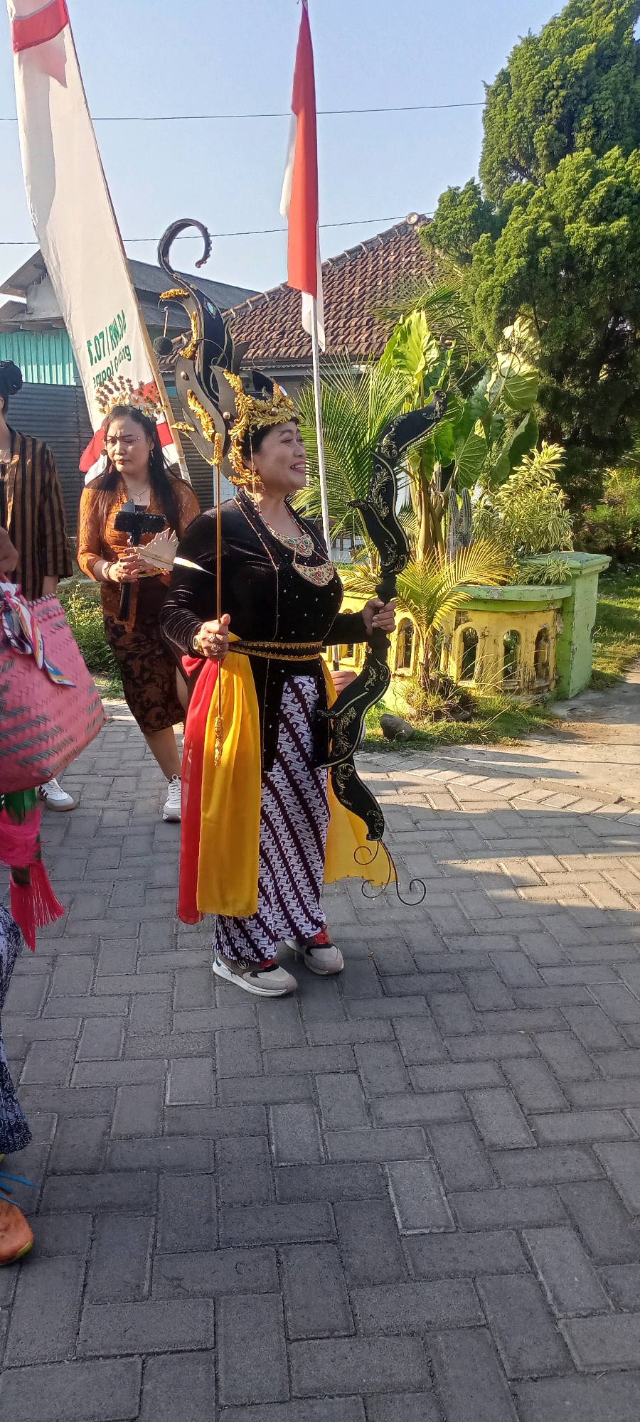 The image depicts a street scene in Gempolklutuk, Indonesia, where a parade or cultural event is underway. In the foreground, a woman dressed in traditional Indonesian attire, including a dark, ornate outfit, a golden headdress, and a patterned sarong, holds a decorative staff with a black, curved element. She is smiling and looking to her right. To her left, another woman in a brown patterned dress and a headpiece holds a small object. Behind them, a tall white flag with red lettering is visible, along with a red and white Indonesian flag. The ground is paved with interlocking grey bricks. In the background, there are houses with tiled roofs and lush green trees and plants, suggesting a semi-rural or suburban setting. The lighting indicates it is daytime, likely late afternoon due to the long shadows. The overall atmosphere appears festive and communal.