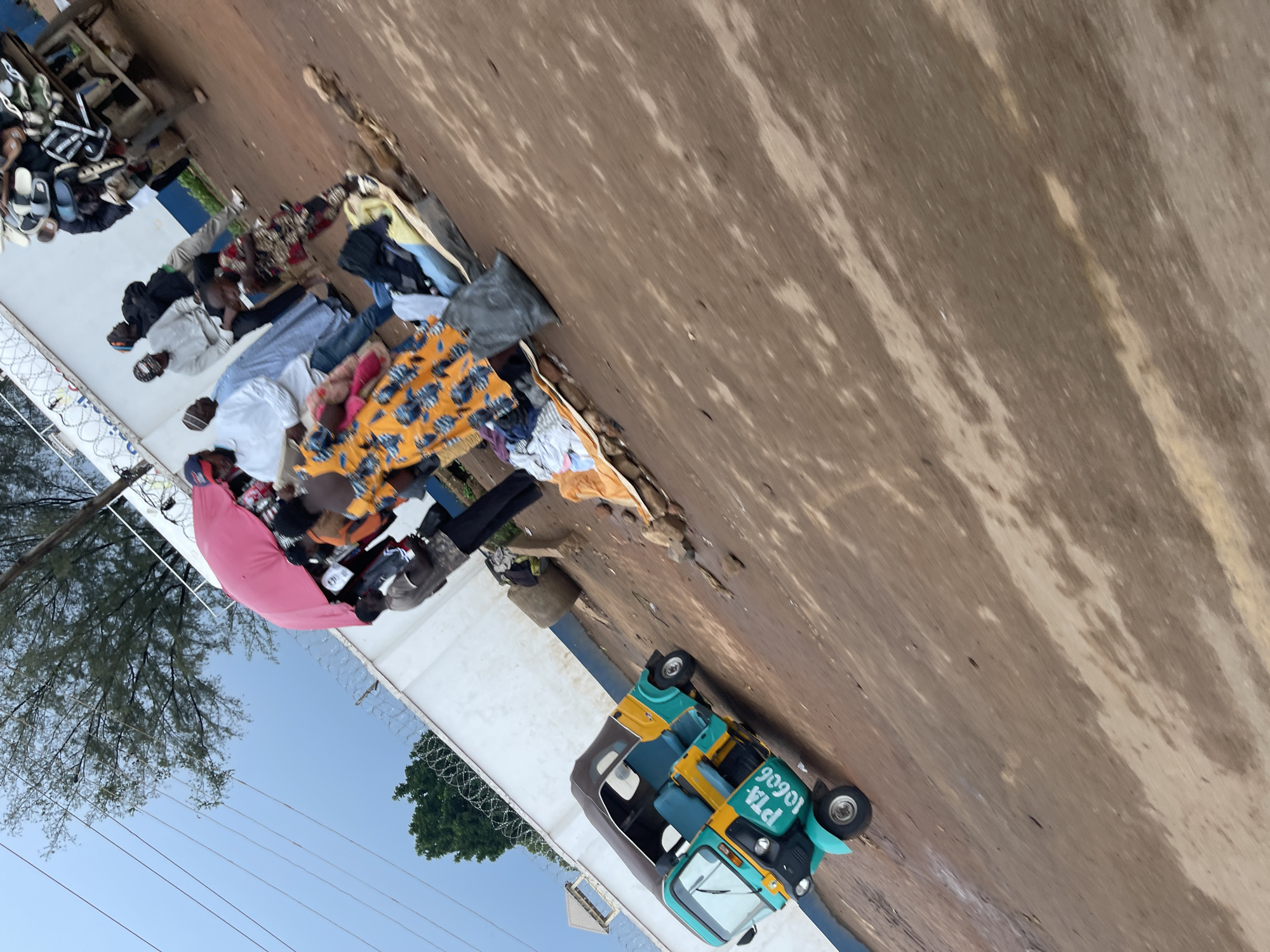 In Rafin Par, Nigeria, a chaotic scene unfolds on a dirt road. A turquoise and yellow auto-rickshaw, displaying the license plate "PTA 10606", is parked at an angle. Several people are gathered around a stall or makeshift market, where clothing and other items are displayed on the ground. A large pink umbrella provides shade over some of the individuals. One person, dressed in a vibrant orange and blue patterned garment, stands out. In the background, a white wall with barbed wire fencing runs along the perimeter, and trees are visible against a cloudy sky. A cart laden with footwear is also present on the right side of the image. The ground is predominantly dirt, with some areas appearing damp.