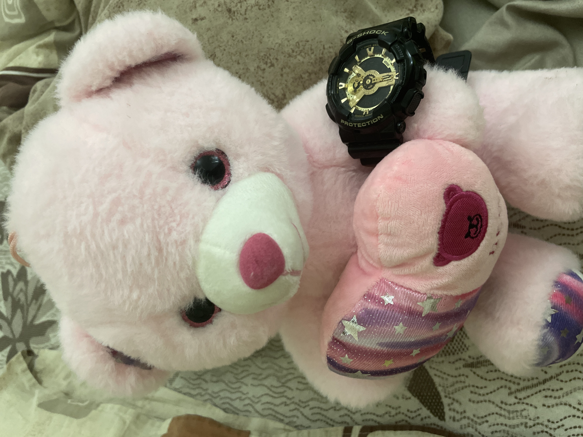 This indoor photograph, presumably taken in Ihtiman, Bulgaria, features a pink stuffed animal lying on patterned bedding. The main subject is a light pink, fluffy teddy bear or similar plush toy with prominent black eyes framed by glittery pink rims. It has a white snout and a small, dark pink nose. One ear is visible at the top left of the frame.
A black G-Shock watch with a black face and striking gold accents is positioned on the stuffed animal's right arm. Visible text on the watch includes "G-SHOCK," "PROTECTION," "1/1000 SEC," "FORWARD," "REVERSE," and the brand name "CASIO."
The stuffed animal displays several distinctive features. Its left foot is adorned with a colorful fabric featuring a blend of purple, pink, and white stripes embellished with silver star patterns. On its other side/arm, there is a rounded dark pink patch with an embroidered symbol resembling the letter "B." The background consists of soft, light-colored bedding with subtle wavy patterns in beige, brown, and white tones. The scene is a still life, characterized by soft, ambient lighting, suggesting a tranquil indoor setting.