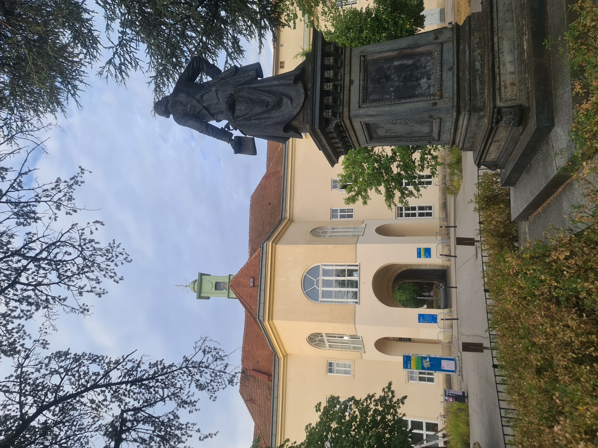 The image depicts a statue of Wolfgang Amadeus Mozart located in the Mozartpark in Vienna, Austria. The statue, made of bronze, is positioned in the foreground, facing away from the viewer. It stands on a large, ornate pedestal. Behind the statue, a light yellow building with a red tile roof is visible. The building features arched entrances and large, arched windows. A green bell tower with a weather vane tops the building. The area around the statue and building is landscaped with trees and bushes, and there is a paved walkway in front of the building. The sky is cloudy.