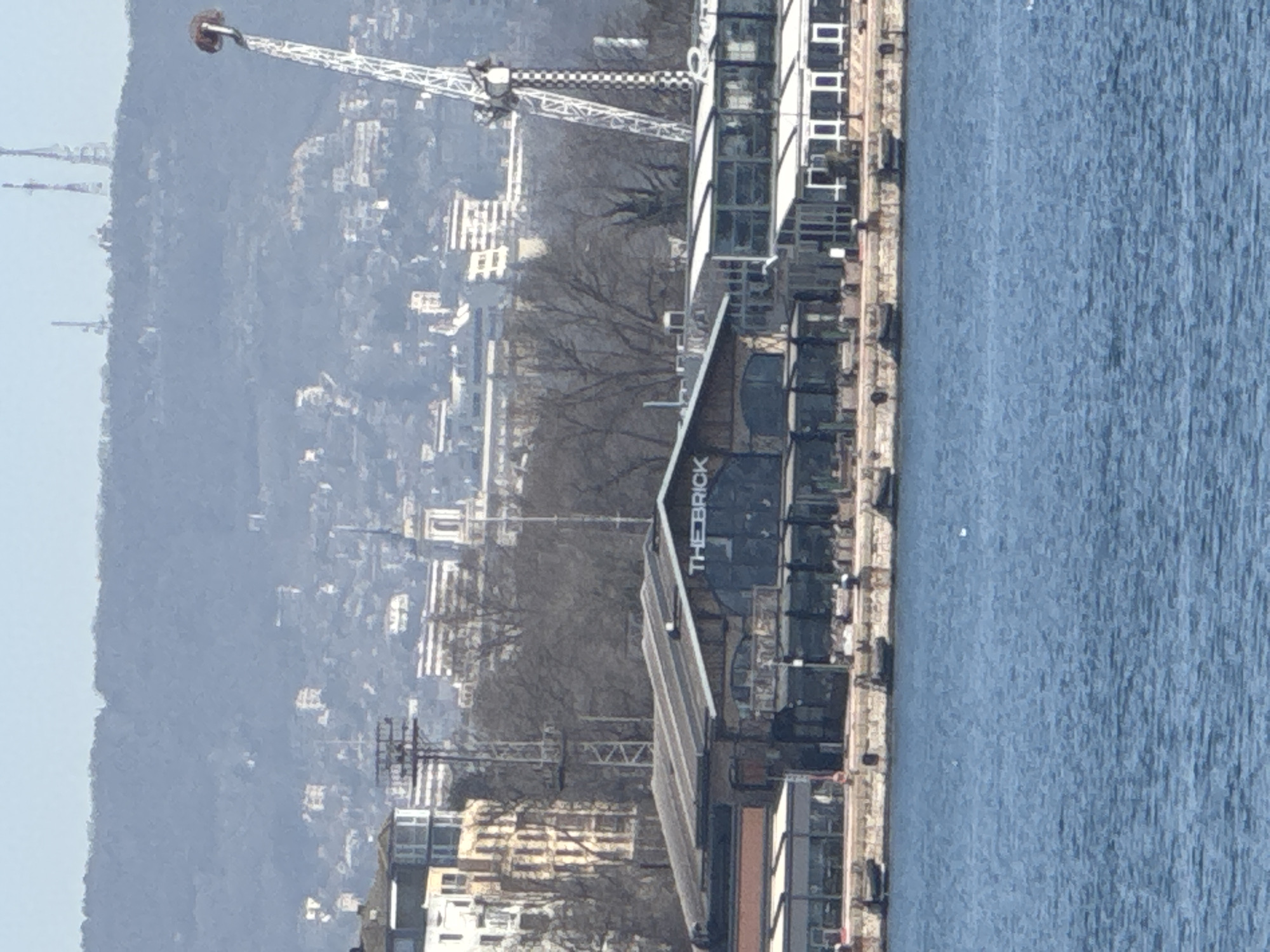 The image shows the building named "The Brick" located on the coast in the city of Varna, Bulgaria. The building is situated next to the water, which occupies a significant portion of the right side of the image. In the background, there is a cityscape with a construction crane visible. The terrain behind the city appears to be hilly and somewhat shrouded in haze. There are no people visible in the image.