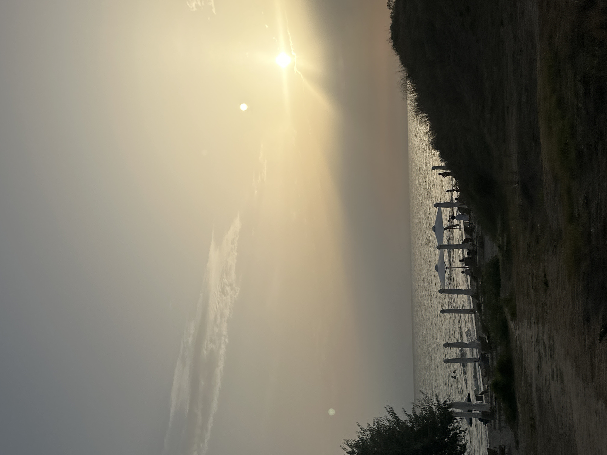 This image depicts a beach scene at dusk in Kassandra Municipal Unit, Greece. The sun is low in the sky, casting a warm, diffused light and creating a hazy atmosphere. The sea is calm with gentle waves rolling towards the shore. Along the coastline, there are several closed beach umbrellas and lounge chairs, suggesting the end of a day at the beach. A few silhouettes of people can be seen in the distance near the water. To the left, the dark silhouette of a tree is visible, and to the right, a grassy dune slopes upwards. The overall impression is one of tranquility and the fading light of day.